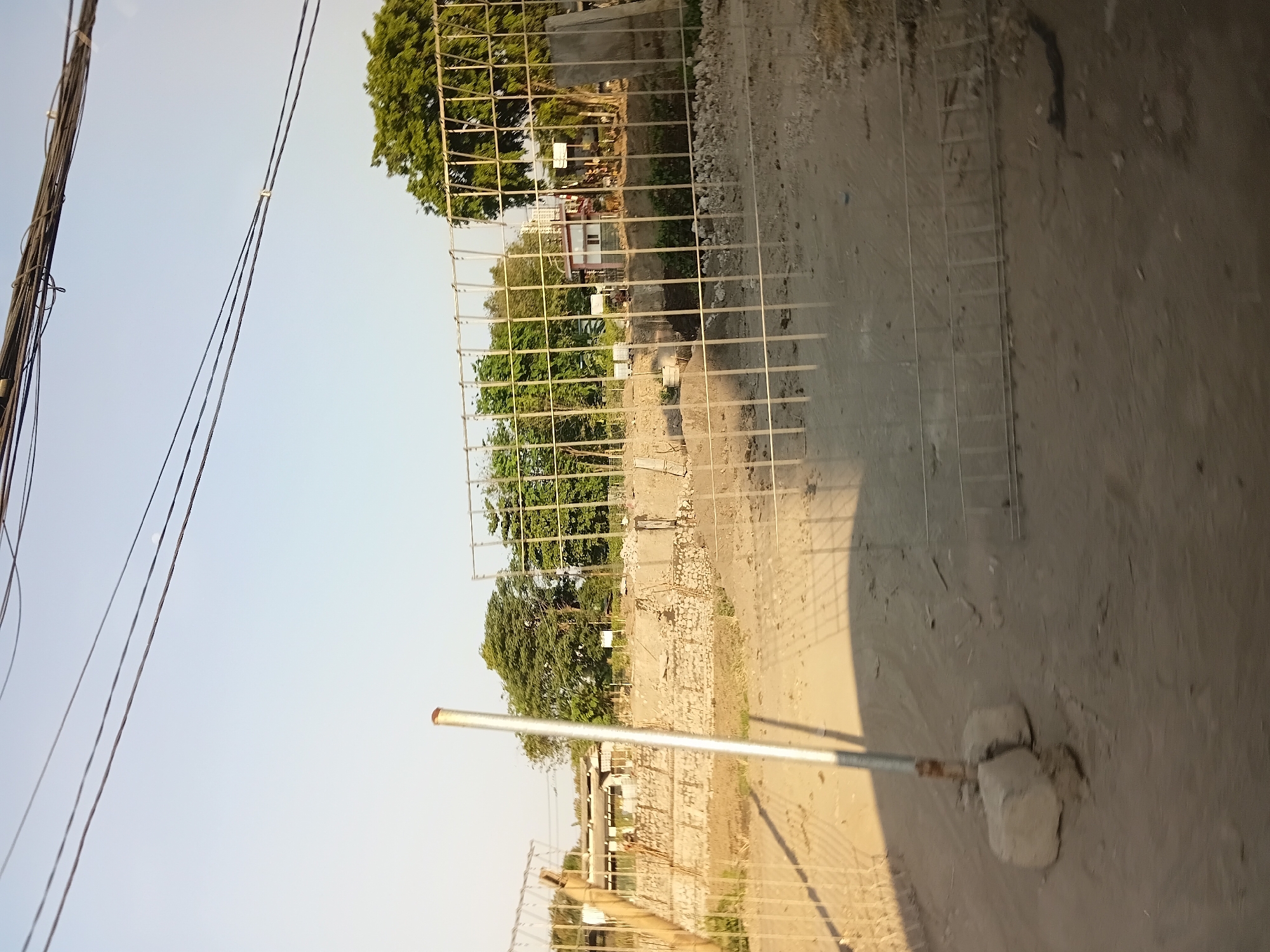 The image shows a construction site with a partially erected metal fence in the foreground. The fence is made of vertical and horizontal metal bars. Behind the fence, there is a disturbed landscape with piles of dirt and stones, suggesting ongoing construction or demolition. In the background, there are several buildings, some appearing to be houses or small commercial structures, surrounded by trees. Overhead, electrical wires are visible against a clear sky. The scene is likely set in Surabaya, Indonesia, given the urban and tropical elements. There are no people clearly visible in the image.