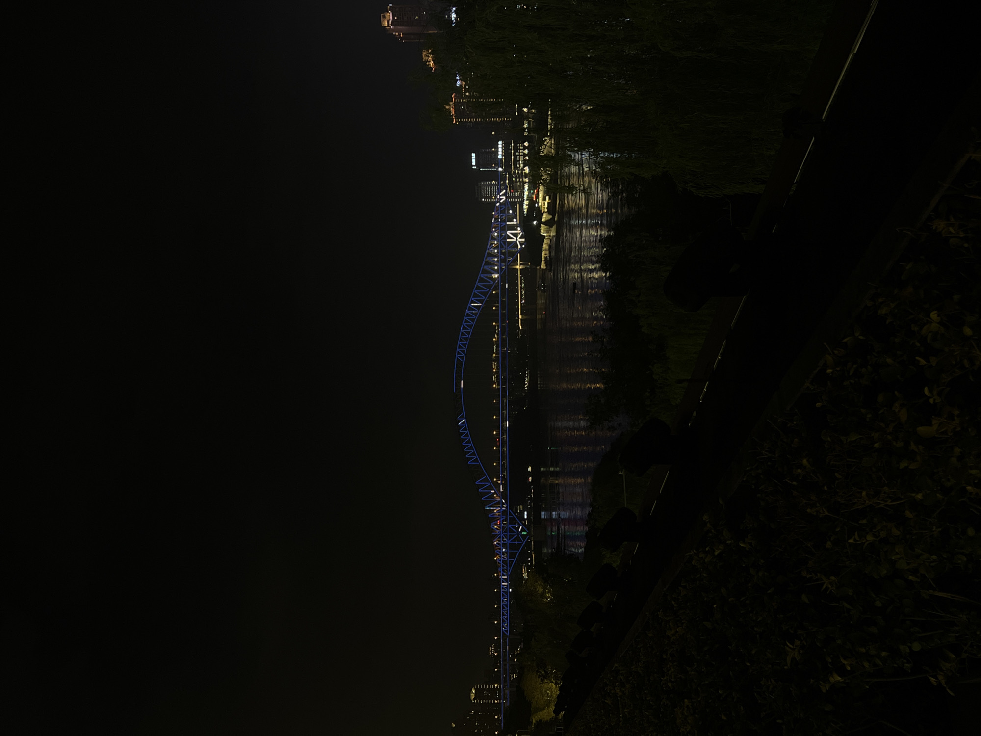 The image depicts a nighttime view of a city skyline with a bridge spanning a river. The bridge is illuminated with blue lights, creating a striking visual against the dark sky. The river below reflects the city lights and the bridge's illumination. In the foreground, there is a dark, landscaped area with some foliage. Several lights are visible on the embankment in the foreground, possibly spotlights. Buildings with illuminated windows are visible on the far bank of the river, suggesting an urban environment. A boat is visible on the river, also illuminated. The overall impression is of a vibrant city at night, with a focus on the illuminated architecture and the river. The location of 江北城街道, China, is known for its waterfront development and illuminated bridges, which are a prominent feature of the cityscape.