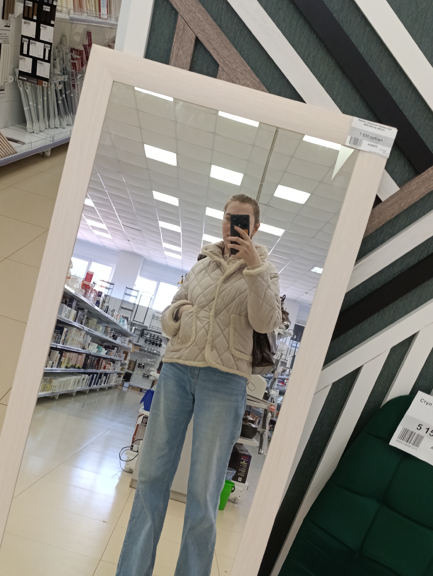 Here is a forensic description of the image:
A person stands in front of a large rectangular mirror with a light-colored frame. The mirror reflects a store aisle lined with shelves full of merchandise. The person in the reflection is holding a phone up, presumably taking a selfie. They are wearing light blue jeans and a beige quilted jacket with a fur-trimmed hood. A brown bag is slung over their shoulder. To the right of the mirror are displays of different wall textures and a green cushioned chair with a price tag of 515. Above the mirror is another price tag stating "7 535 руб/шт". The overall environment appears to be a retail store, likely in the city of Chaikovsky, Russia, based on the currency symbol on the price tag. The store has bright overhead lighting.