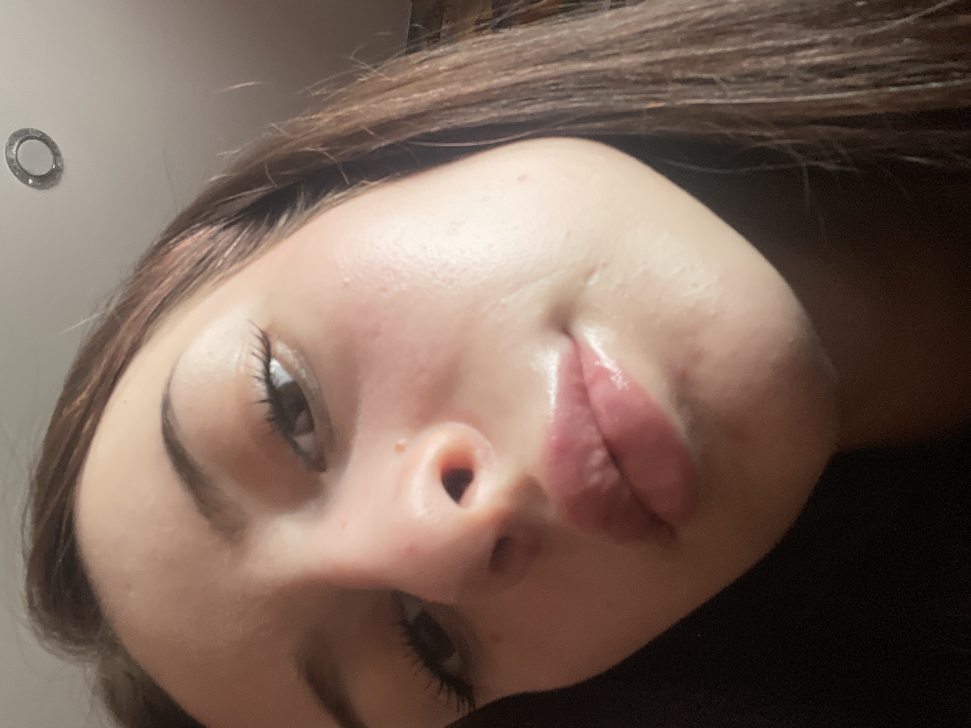 The image is a close-up, low-angle selfie of a young woman's face. Her hair is dark brown and falls loosely around her face. Her eyebrows are well-defined, and her eyelashes are long and dark. Her skin appears smooth and slightly dewy, with some subtle redness on her cheeks. Her lips are full and have a glossy pink appearance. The lighting suggests an indoor setting, possibly a bedroom, with a ceiling light fixture visible in the upper right corner. There is no identifiable background that would indicate a specific location within the city of Medvevedo, Russia. The focus is entirely on her face.