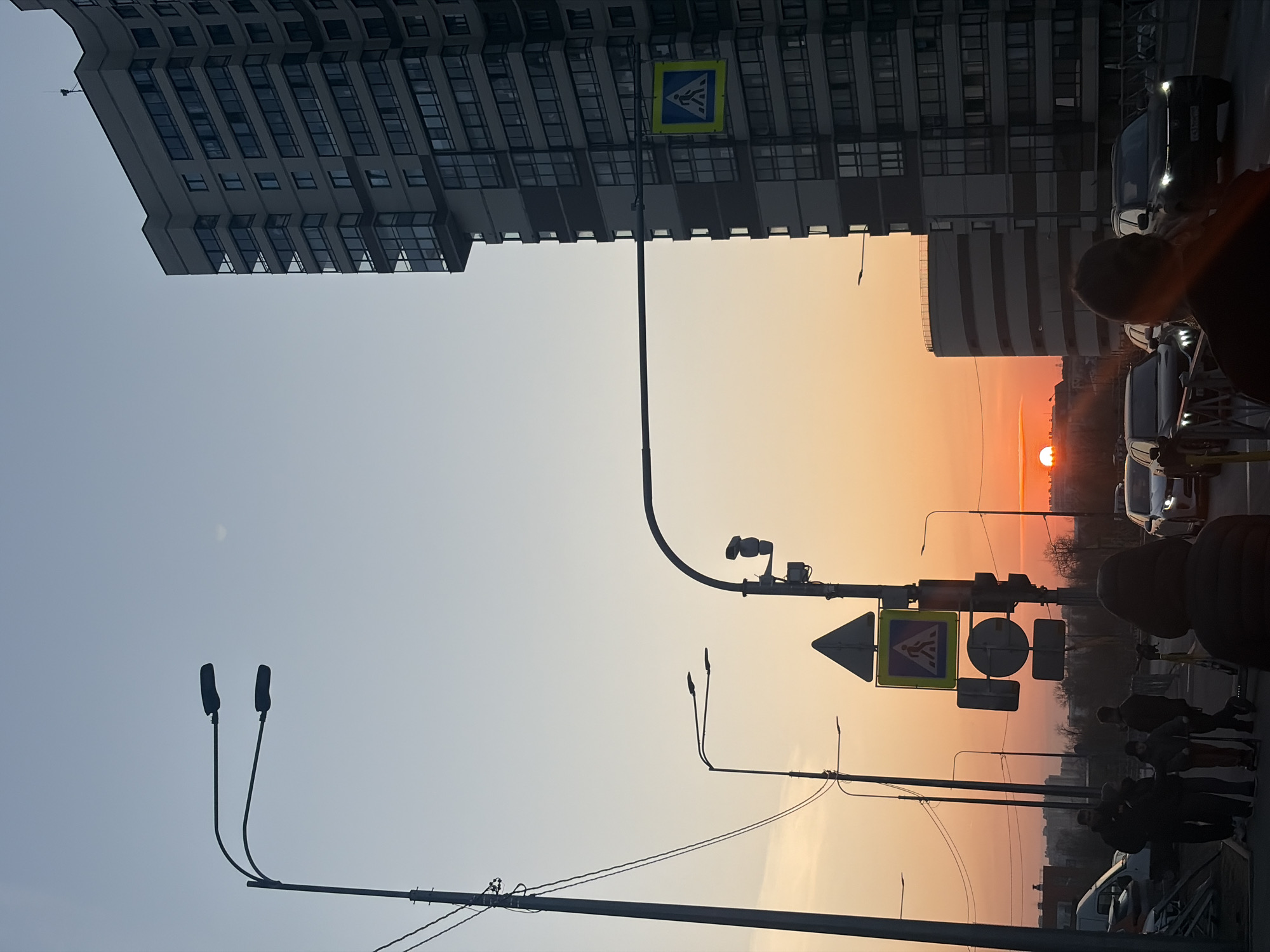 Here's a description of the image:
The image depicts a street scene in Saint Petersburg, Russia, during a sunset. The sky is a gradient of colors, transitioning from blue to orange as the sun sets on the horizon. Several buildings are visible, including a tall residential building and a rounded building with a unique architectural design. Street infrastructure includes pedestrian crossing signs, traffic lights, and lamp posts. There are vehicles, including cars and vans, along the street. The sidewalk is lined with people and a few barrels, indicating pedestrian traffic and possibly construction or road maintenance.