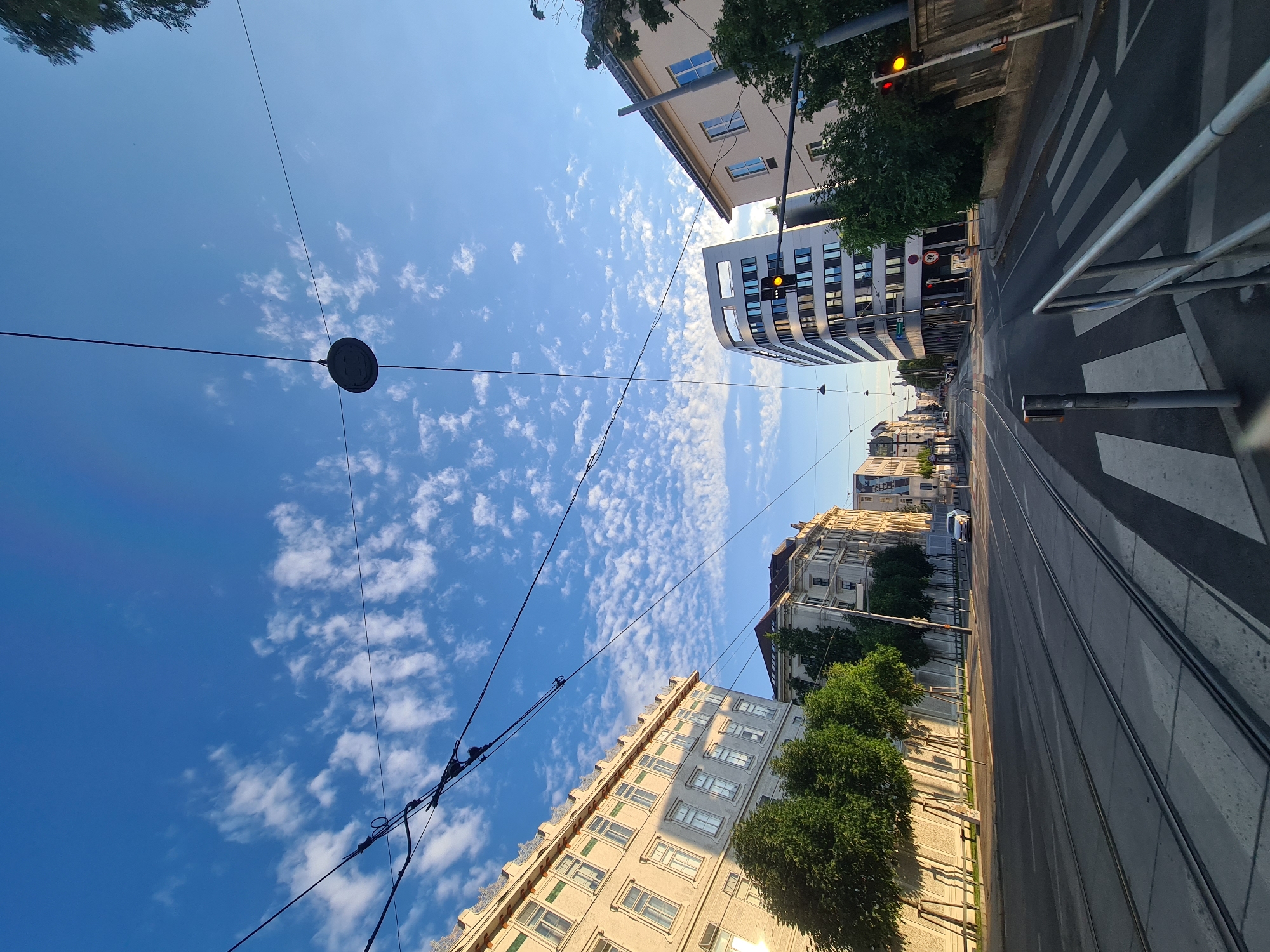 The image captures a street scene in Vienna, Austria, with a perspective looking down a wide road featuring tram tracks. The sky is a clear blue with scattered white clouds, suggesting it is daytime.
On the left side of the street, a light-colored building with multiple windows and decorative elements along its roofline is visible. Adjacent to it are several mature trees lining the sidewalk. Further down the street, another similar-looking building can be seen.
On the right side of the street, a modern, curved building with many windows stands prominently. In front of it, a traffic light shows a yellow signal. A pedestrian crossing with white stripes is marked on the road, and a metal railing runs along the sidewalk.
The road itself is paved and shows visible tram tracks. A white car is seen in the distance, driving away from the camera. Overhead, numerous black wires crisscross the sky, connected to poles and a hanging circular fixture, characteristic of a tram network. The overall impression is a well-maintained urban environment, likely in a business or residential district of Vienna.