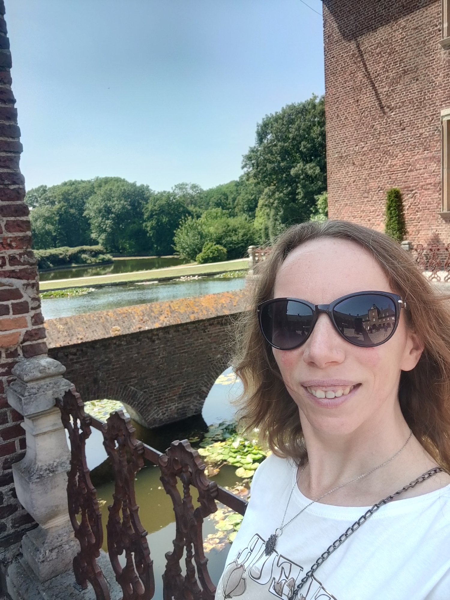 The image is a selfie taken by a woman in front of a body of water and a brick building. The woman is wearing sunglasses, a white t-shirt, and a silver necklace. In the background, there is a bridge made of brick, trees, and a clear blue sky. The reflection in the woman's sunglasses shows a building that appears to be a castle or a large historical building. The scene is likely outdoors, possibly in a park or garden area. The presence of a brick structure resembling a castle and the mention of Isselburg, Germany suggests the location could be a historical site in or around that city.