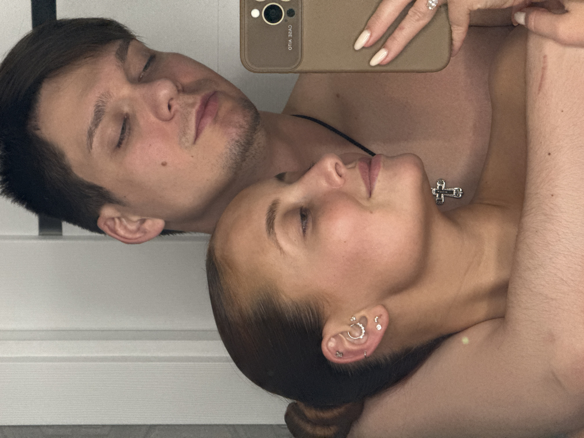 A selfie shows two light-skinned individuals, a male and a female, lying together. The male, on the left, has short dark hair and closed eyes. The female, on the right, also has closed eyes and multiple piercings in her left ear. A cross necklace is visible on her chest. The phone used to take the photo is visible at the top center, held by a hand with manicured nails and a ring. The location, Chaikovsky, Russia, cannot be verified based on the visual information provided.