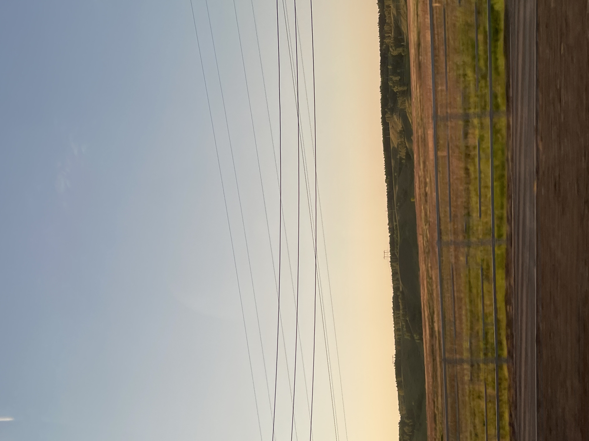 The image, correctly oriented, presents a broad panoramic view of a rural landscape under an expansive sky, captured during either sunrise or sunset. The sky dominates the upper half, displaying a horizontal gradient from a clear, light blue on the left to a warm, golden-orange hue on the right, characteristic of a low sun angle. Faint, wispy clouds are visible in the lighter blue portion of the sky.
Several dark, parallel power lines stretch horizontally across the sky, silhouetted against the bright background. These high-tension lines serve as a prominent man-made element within the natural setting.
The horizon is defined by a distant, dark green band of forest, indicating a continuous tree line or wooded hills. Below the horizon, the landscape consists of agricultural fields. These fields are marked by long, parallel strips of differing colors and textures, featuring sections of muted brown earth alternating with vibrant green vegetation, suggesting varied crops or cultivated land.
In the far distance, subtly visible on the right side of the horizon, is the silhouette of a power transmission tower. A vertical wooden structure, likely a fence or barrier, partially appears along the extreme right edge of the frame.
The scene is devoid of human or animal activity, offering a tranquil and vast depiction of the countryside. The warm lighting and open fields convey a sense of peacefulness and natural grandeur, consistent with the rural environment of Элита, Russia. No text is visible in the image.