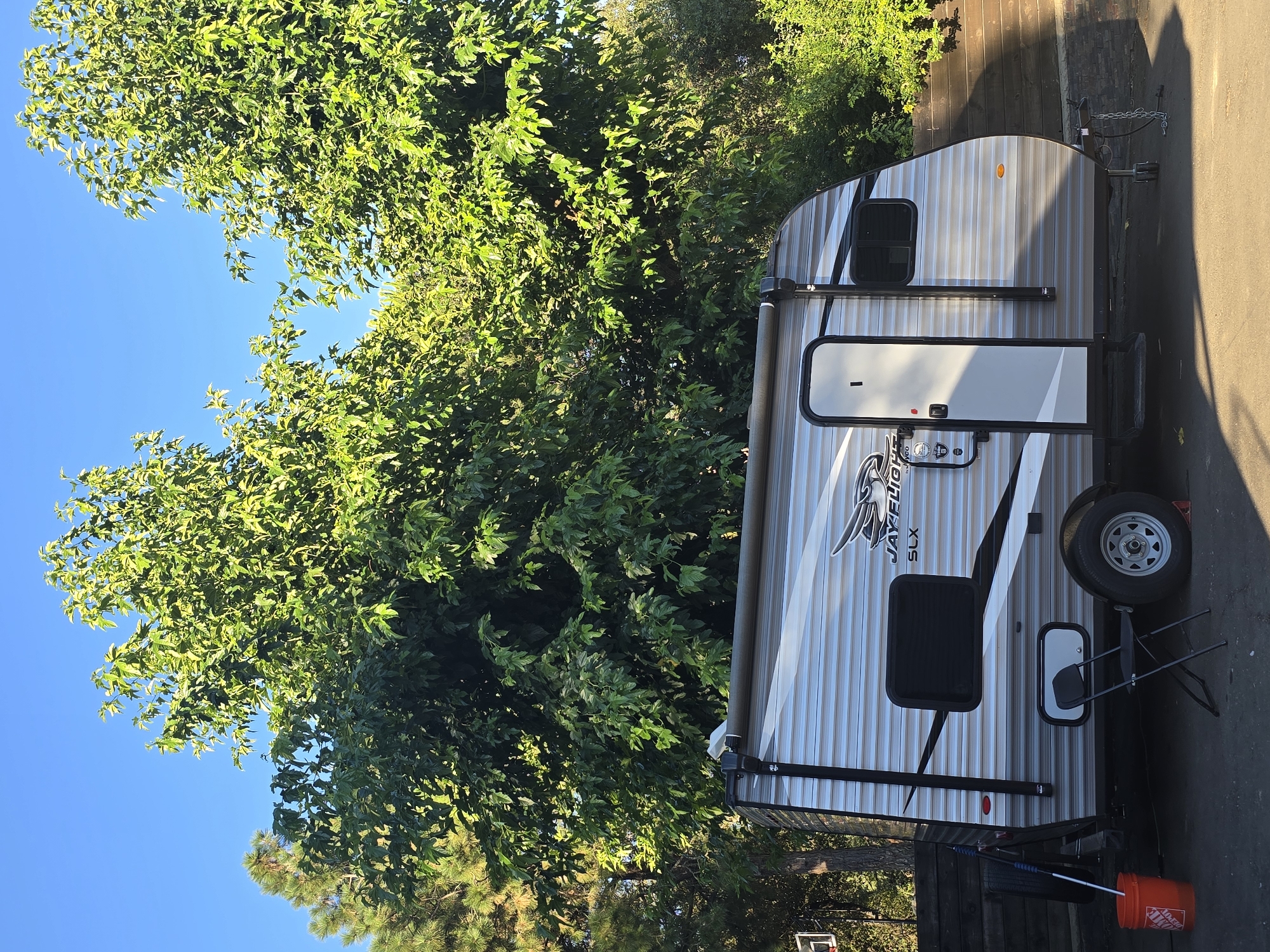 A travel trailer is parked outdoors in front of a wall of trees. The trailer has a white door with a silver handle and a window next to it. The side of the trailer has a silver and white striped design with a black awning. There are two windows on the side of the trailer, one is black and the other is white. The trailer has a black wheel with silver trim. To the right of the trailer is a black folding chair. In front of the trailer is an orange bucket and a black mop. The trailer has "JAY FLIGHT SLX" written on the side. The image is taken in daylight with a clear blue sky and bright green trees. The sun is casting shadows on the ground, indicating it is likely late afternoon. The setting appears to be a residential area or campground.