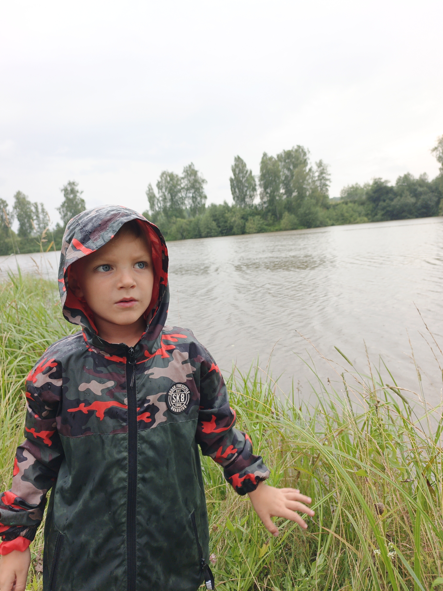 A young boy wearing a camouflage hooded jacket stands in tall grass near a body of water. The boy's face is turned to the right, with his mouth slightly open. The jacket has a red lining and a circular patch on the chest with text. The water appears calm, with trees lining the opposite bank. The sky is overcast. The location is identified as Urtiushka, Russia.