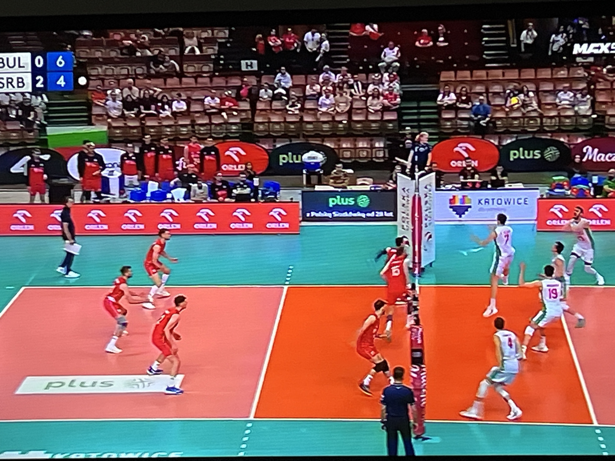 The image shows a volleyball match in progress. On the left side of the net, players wearing red and white uniforms are preparing for a play. A person in a dark uniform stands near the net. On the right side, players in light blue and white uniforms are positioned defensively. The scoreboard indicates Bulgaria (BUL) has 0 points and 6 in this set. Serbia (SRB) has 2 points and 4 in this set. Spectators are visible in the stands behind the court. Advertising is visible on the court floor and backdrop, featuring brands like "plus" and "ORLEN". The backdrop indicates the match is held in Katowice. The image gives no indication of the location being in the city of Gabrovo, Bulgaria.