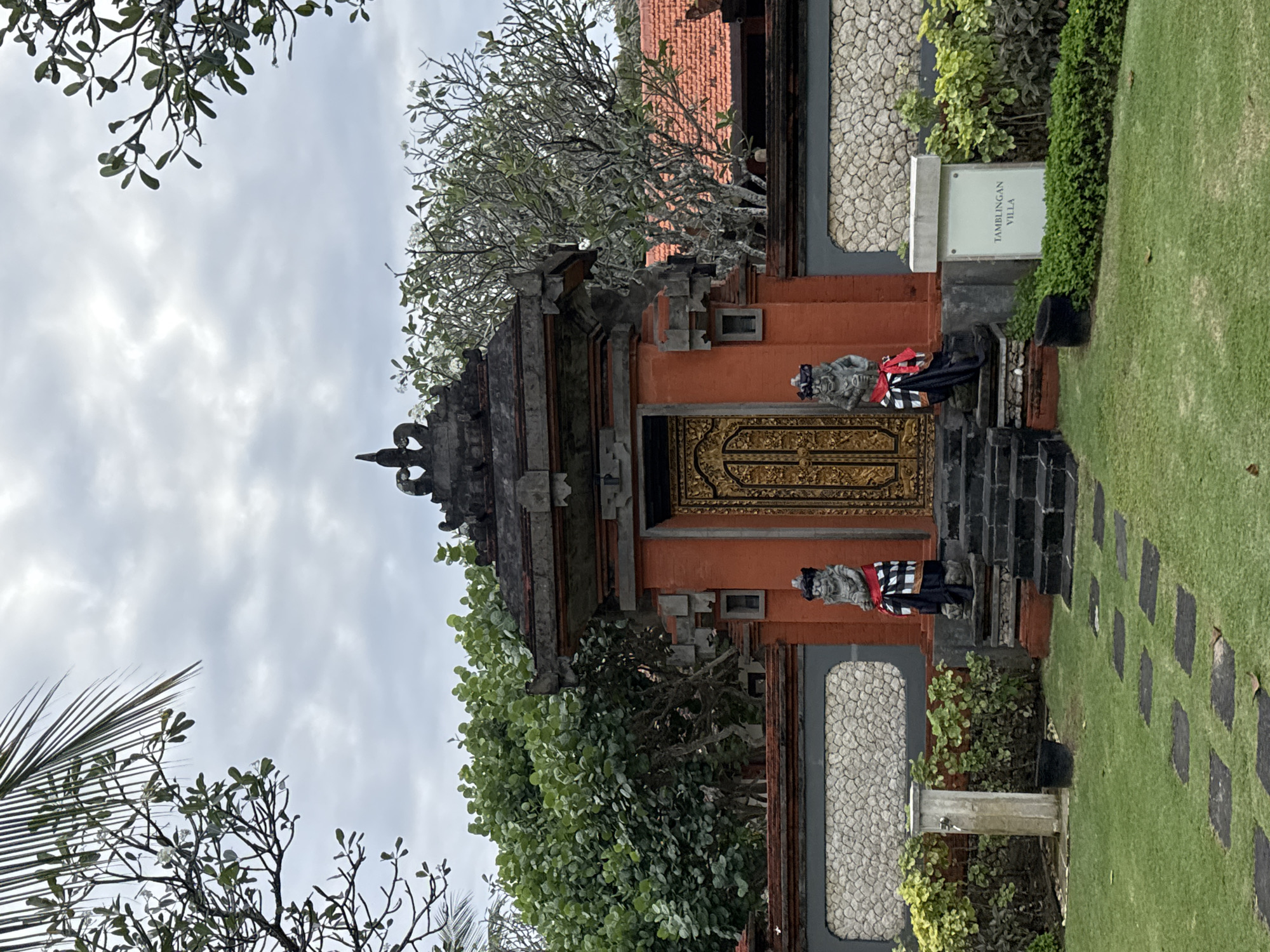 The image shows the entrance to Tamblingan Villa in Kuta Selatan, Indonesia. A traditional Balinese gate, painted orange with ornate gold doors, is centrally located. Flanking the gate are two stone statues dressed in traditional black and white checkered sarongs with red sashes. Stone steps lead up to the gate. To the left of the entrance, a stone pillar with a faucet stands in a manicured lawn, next to a dark bucket. To the right of the entrance, a sign reads "TAMBLINGAN VILLA". The entrance is surrounded by a stone wall and lush green foliage. Palm fronds are visible in the upper left corner, and a cloudy sky completes the scene.