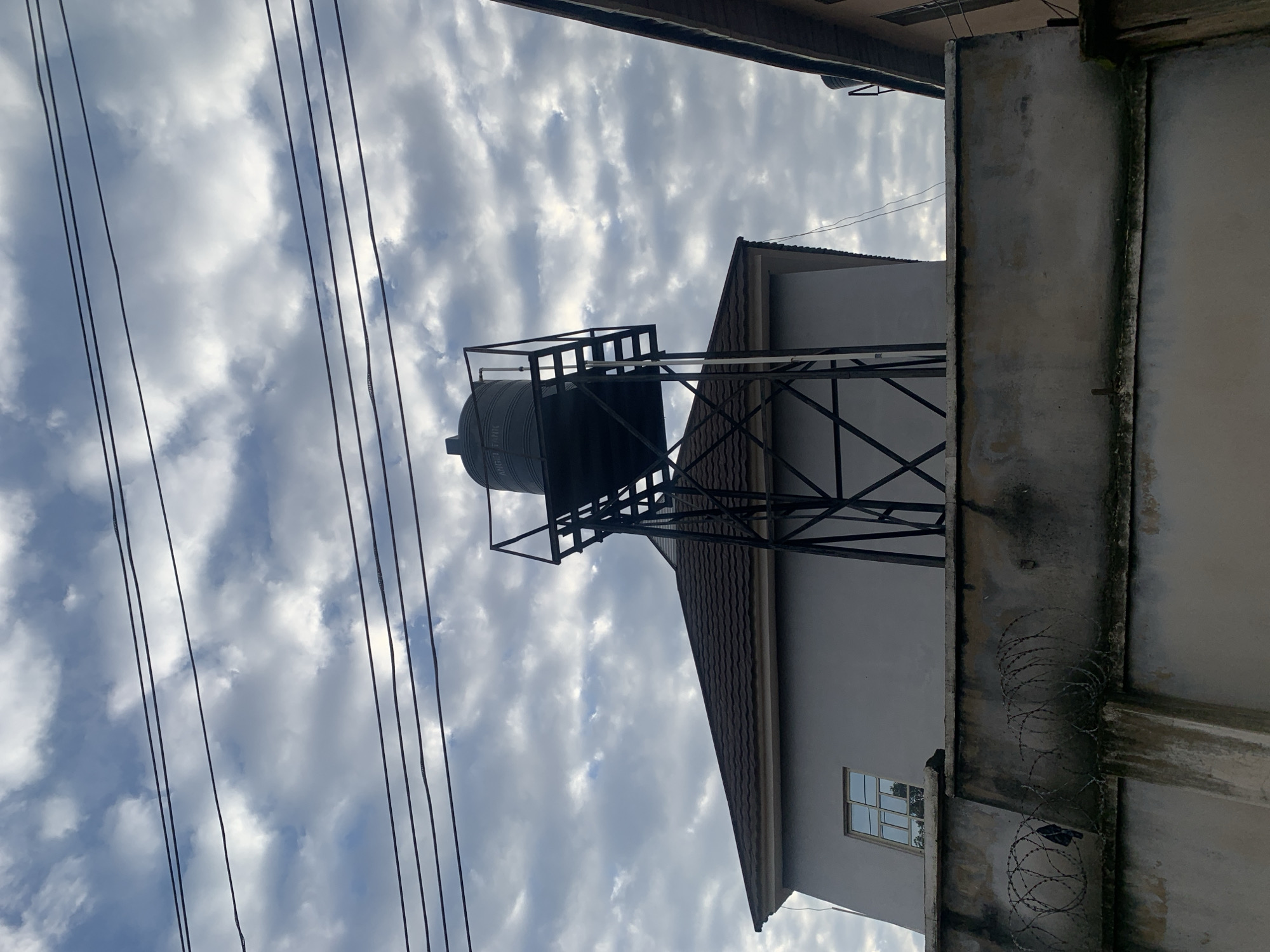 The image shows a water tank elevated on a metal stand above a residential building. The building has a dark-tiled roof and a light-colored exterior wall with a window. In front of the building is a concrete wall topped with barbed wire. Several power lines are visible against a cloudy sky. The scene appears to be in Rafin Par, Nigeria.