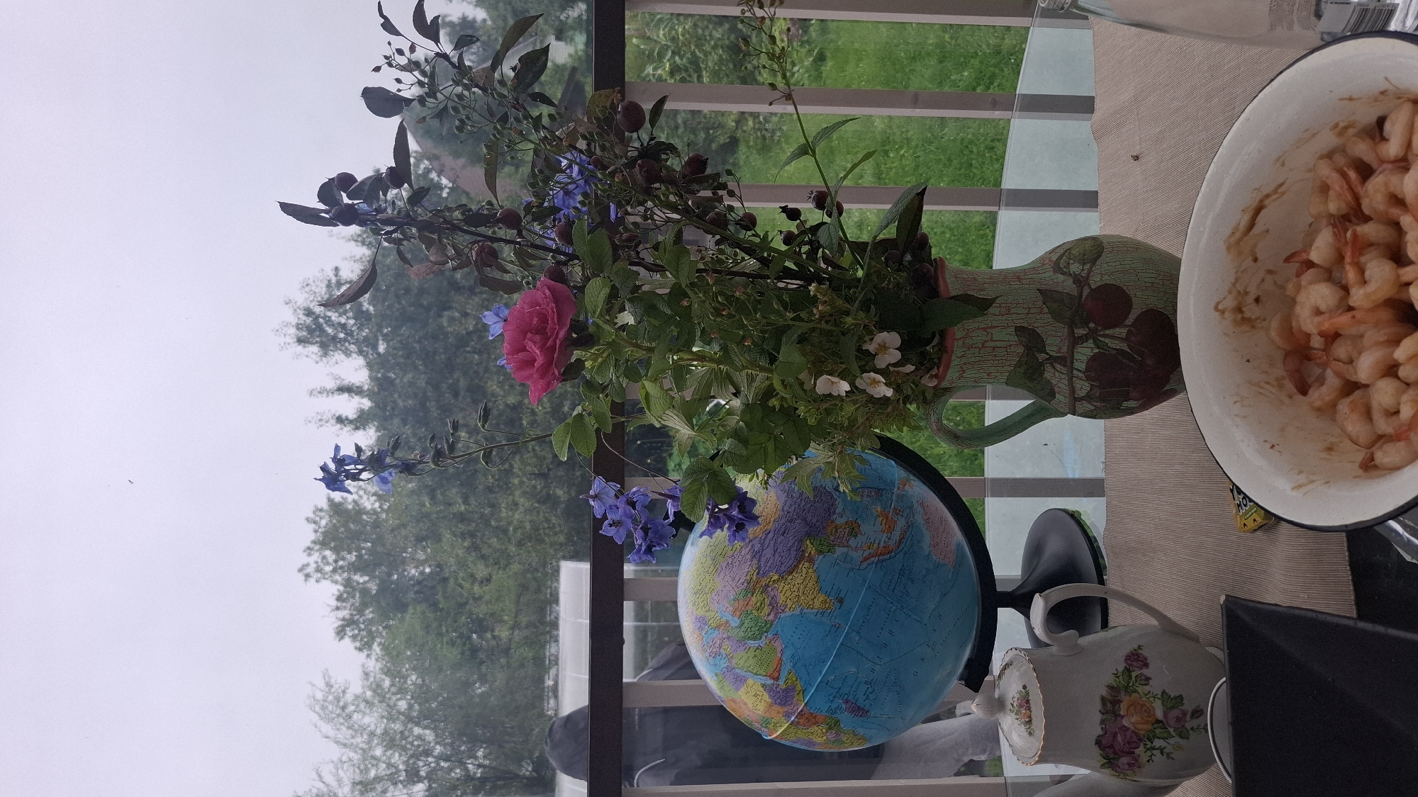 This image depicts an outdoor setting on a table, likely a balcony or patio, with a backdrop of trees and an overcast sky. The foreground features a collection of objects: a globe with a black stand, a decorative teapot with floral motifs, a vase filled with a bouquet of flowers and berries, and a bowl containing cooked shrimp. A person wearing a dark hooded jacket is partially visible in the background, standing near a railing. The overall scene suggests a casual gathering or moment of contemplation in a natural, possibly rural, environment. The location is stated to be Орбита Снт (терпигорьево), Russia.