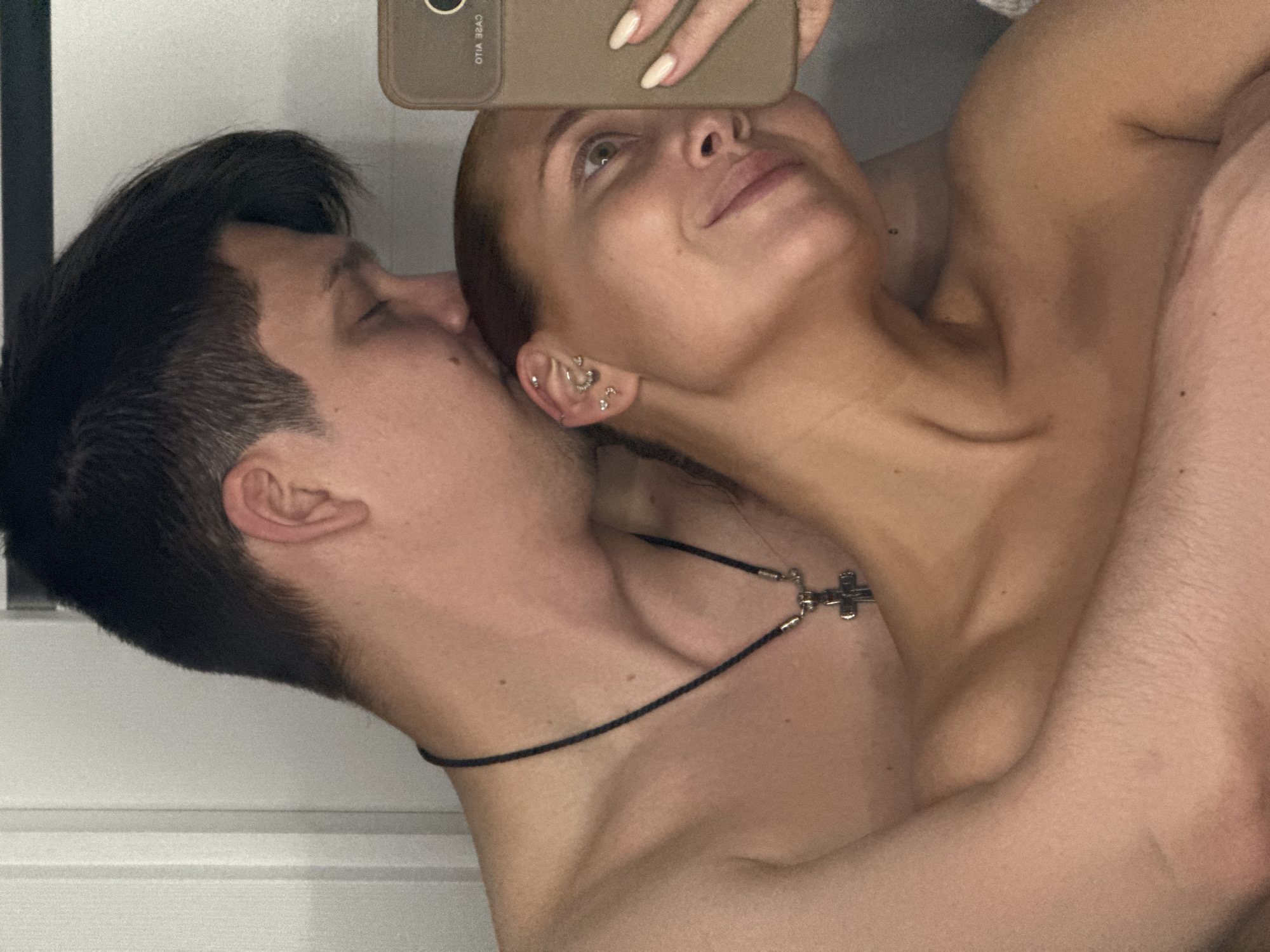 A shirtless man with dark hair and a shirtless woman with her hair pulled back are in close proximity. She is looking upwards and holding a phone in front of her face. She has multiple piercings in her left ear. A black cord with a metal cross is visible around the man's neck. The background is a plain, light-colored wall. The location is not discernible.