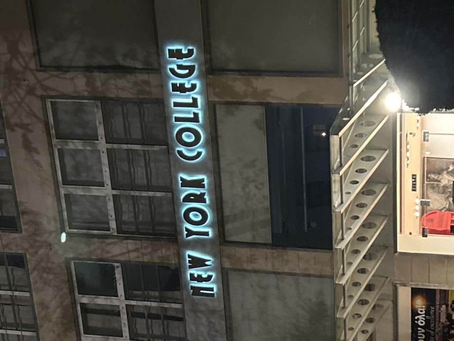 The image captures the exterior of a building at night, viewed from a slightly upward angle. A prominent, vertically oriented, illuminated sign in glowing light-blue letters reads "NEW YORK COLLEGE". This sign is affixed to a dark grey vertical panel on the building's facade.
The building itself features multiple rows of square or rectangular windows with dark frames, reflecting the nighttime darkness and some ambient light. The facade appears to be constructed from light-colored, possibly textured, panels, showing some signs of age or weathering.
To the right of the main building section with the sign, there is a striking architectural element, possibly an external staircase or decorative screening. This structure is composed of repeating triangular segments, each perforated with several circular holes. It is also lit by several bright lights, creating a luminous effect and casting light onto the adjacent surfaces.
At the bottom right corner, another section of a structure or a billboard is visible, displaying text. Part of this text is in Greek, appearing to end with "...ουν όλα!", followed by English text that includes "...of excellence". This suggests an advertising slogan or motto for the college. The overall scene is dark, highlighting the illuminated elements against the night sky, indicating a commercial or educational establishment in an urban setting. The location context indicates this is a branch of New York College in Kallithea, Greece.