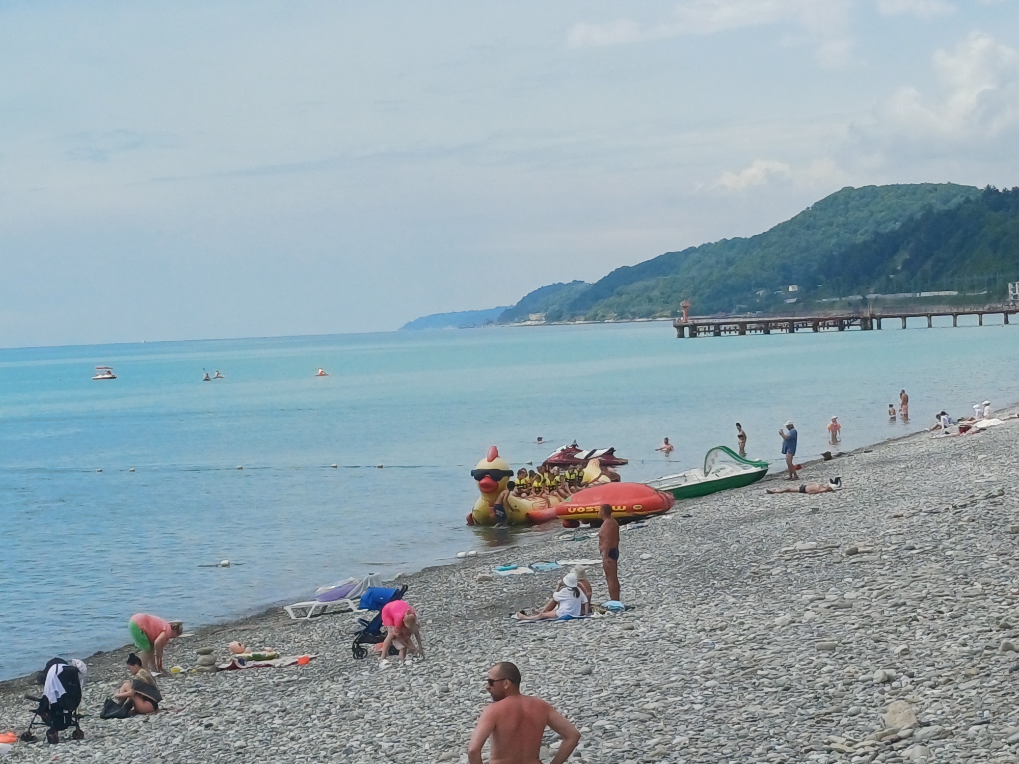 This is a wide-angle shot of a pebble beach and the Black Sea coast in Sochi, Russia. In the foreground, a man wearing sunglasses and no shirt stands facing away from the camera. To his left, a group of people are scattered across the beach. A child in a pink shirt is playing near a stroller. Further back, a woman in a pink top and green shorts bends over. To her right, a woman with dark hair sits on a towel. More people are visible on the beach and wading in the water. In the sea, a large inflatable duck-shaped watercraft with a group of people on it is partially submerged. Several other inflatable watercraft are nearby. The sea is a clear blue, and a long pier stretches into the water in the distance, with a lighthouse at its end. Lush green hills rise behind the coast. The sky is a pale blue with scattered clouds.