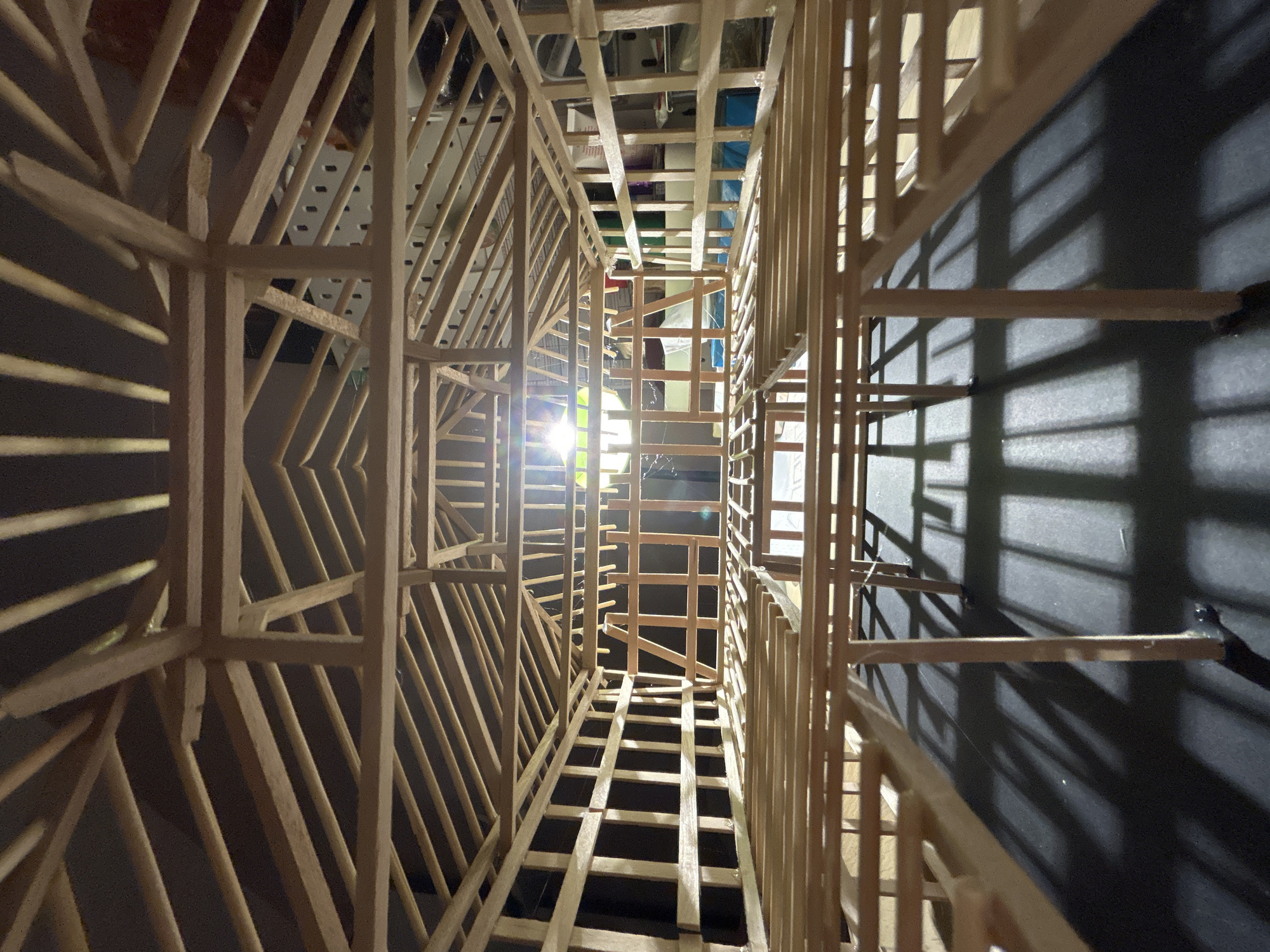 The image shows a low-angle, close-up view looking up through the interior wooden framework of a model house. The model is constructed from thin wooden strips and resembles a skeletal structure with multiple floors and a gabled roof. The lighting suggests an indoor setting, with a bright light source illuminating the central portion of the model, casting long shadows on the dark surface below. The model is unfinished and appears to be in the process of construction. There are no people or animals visible in the image, nor any indication of a specific location in Sofia, Bulgaria.