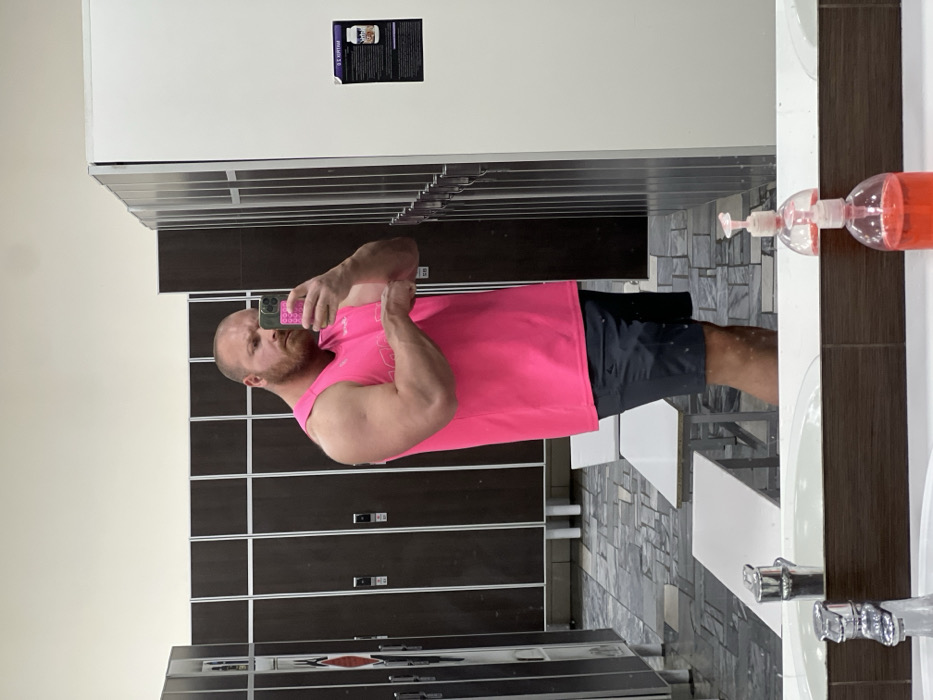 A muscular, bald man with a reddish-brown beard, wearing a bright pink sleeveless top and dark shorts, is captured in a mirror selfie within what is described as a locker room in Chaikovsky, Russia. The man holds a smartphone in his right hand, facing the mirror, while his left arm is bent. The background consists of rows of dark brown lockers with white frames and handles, extending along the walls. Two white rectangular benches with light-colored metal legs are positioned on the multi-toned grey and black tiled floor. The upper walls are painted a light, neutral color. To the right, a white locker panel displays a dark sign with white text and a product image. In the foreground, below the mirror, a white sink with two chrome faucets is visible on a brown counter, reflecting two clear pump bottles containing orange liquid.