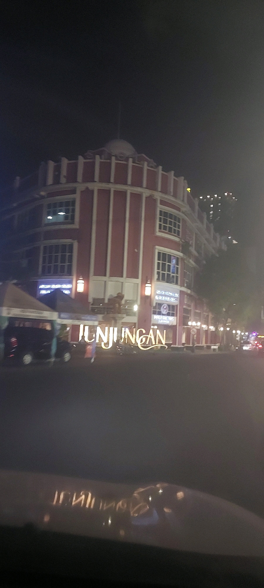 A night shot of a building with red and white accents, featuring a dome-like structure on top. In front of the building, large, illuminated letters spell out "Tunjungan". There is a dark-colored van parked to the left, and a few people can be seen in the vicinity. The image is slightly blurred, suggesting it was taken from a moving vehicle. The "Tunjungan" signage indicates the scene is located in the Tunjungan area of Surabaya, Indonesia.