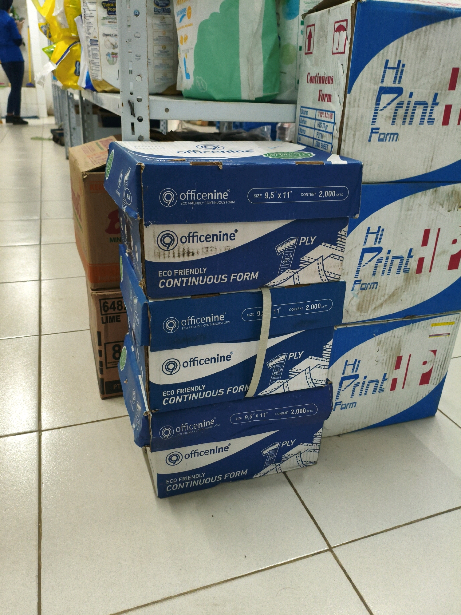 A stack of office nine brand continuous form paper boxes is the primary subject of this image, taken in what appears to be a commercial or storage area. The boxes are primarily blue with white text and logos. One box displays "OfficeNine ECO FRIENDLY CONTINUOUS FORM" and "SIZE 9.5\" x 11\" CONTENT 2,000 SETS". Two larger, partially visible boxes, branded "Hi Print Form", are positioned to the right of the stack. These larger boxes are white with blue and red lettering and graphics, including an umbrella symbol. In the background, shelves are stocked with various white and green bags or packaging. A person wearing a blue shirt and dark pants is visible in the far left background, walking away from the camera. The floor is tiled with a beige and white pattern. The lighting is bright, suggesting an indoor environment. The location is identified as Sumberejo Kulon, Indonesia.
