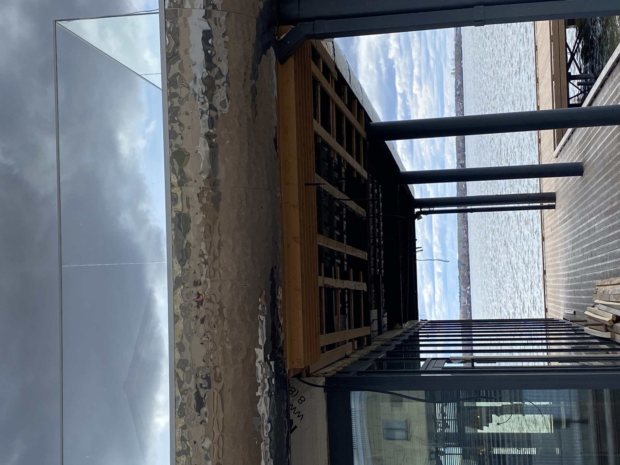 This outdoor image depicts a construction or renovation site, likely in Votkinsk, Russia, given the context. The primary focus is on architectural elements: a reflective surface, perhaps a mirrored or metallic facade; exposed wooden framing; metal support beams; a brick-laid patio or walkway; and windows with visible reflections. The background reveals a body of water with a distant shoreline under a partly cloudy sky.