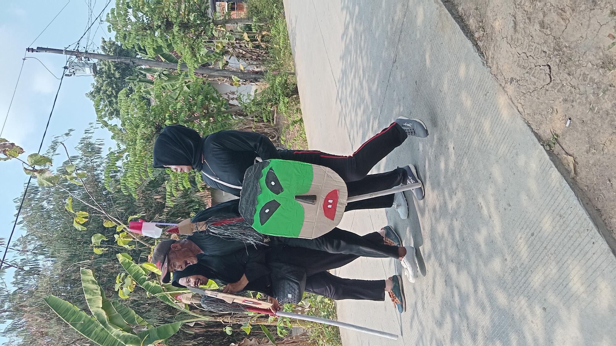 Three individuals are walking on a paved road in a rural or suburban area, possibly Belor, Indonesia, during daylight. They are all dressed in black clothing, and two of them are wearing hijabs. The person in the center, an older man wearing a cap, is yelling or singing. He is holding a stick with a small Indonesian flag attached to it. The person to his left, a woman, is also holding a stick. The person to the right, a woman, is carrying a large, flat, green mask with black hair, white eyes, and red lips. The mask is attached to a stick. The background shows lush green trees and vegetation, as well as a utility pole and some buildings. The road is made of concrete with visible expansion joints. The overall atmosphere suggests a festive or celebratory event, possibly a parade or procession.