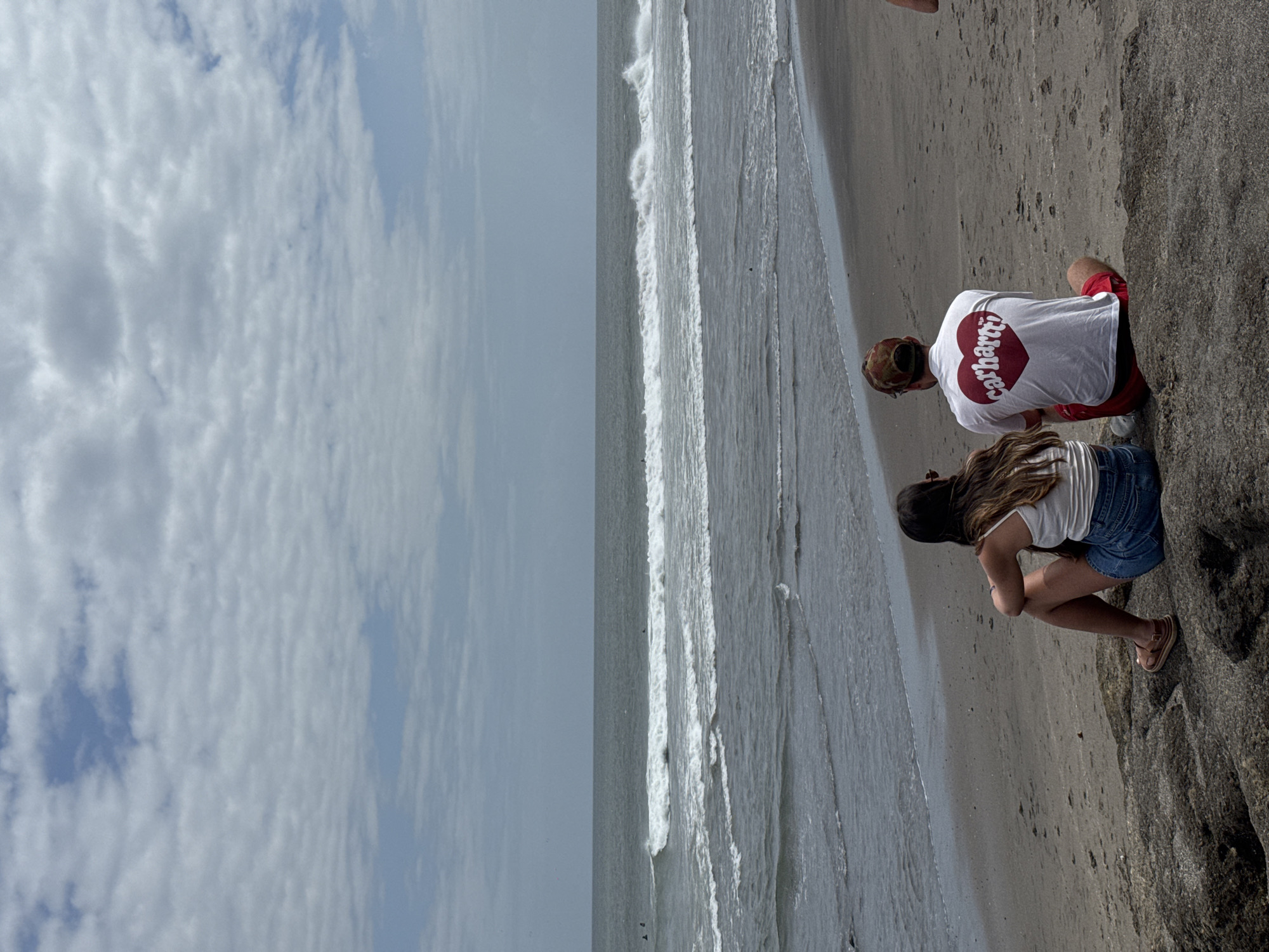The image shows a coastal scene, likely a beach, with two people crouching near the shoreline. The sky is overcast with patterned clouds. The water has small waves, and the beach transitions into a rocky area. One individual wears a white t-shirt with a red heart logo, red shorts and has short brown hair, while the other individual wears a white tank top, denim shorts, and has long brown hair. They appear to be interacting with something on the sand or near the rocks. There is no visual confirmation of being in North Kuta, Indonesia.
