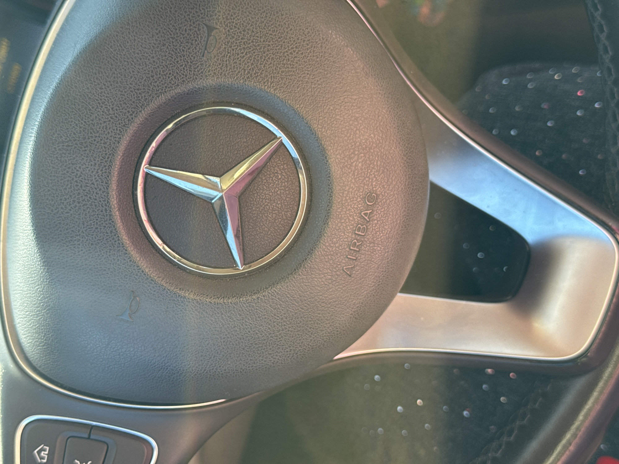 This is a close-up, eye-level, indoor shot of a Mercedes-Benz steering wheel, likely in a car. The prominent subject is the iconic Mercedes-Benz three-pointed star logo, centered on the airbag cover. The steering wheel is made of dark, textured material, possibly leather. To the right of the logo, the word "AIRBAG" is embossed into the material. There are subtle symbols of a horn on either side of the logo, indicating the horn buttons. Below the main steering wheel area, a portion of the vehicle's controls is visible, including buttons with embossed symbols. The lighting suggests it's daytime, with reflections of light and possibly the interior of the car visible on the metallic trim of the steering wheel and the dashboard area. The overall scene is clean and focused on the car's interior details. There are no people visible in the frame. The context provided places this image in Saint Petersburg, Russia.