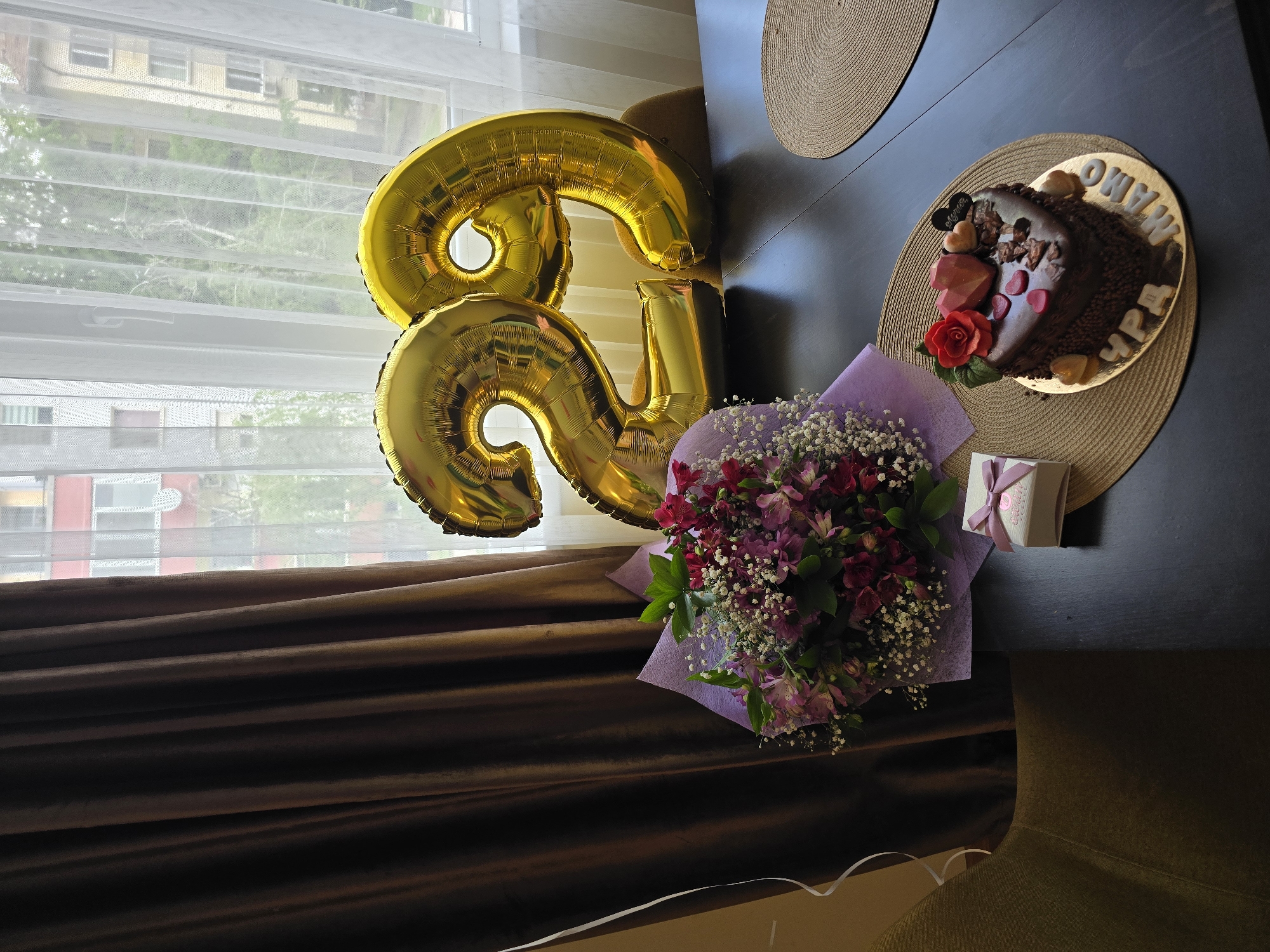 This media file captures an indoor scene depicting a celebration, likely a birthday, next to a window.
The main subjects are two large, gold foil balloons forming the number "83," a heart-shaped chocolate cake, a bouquet of flowers, and a small gift box. The "83" balloons are prominently displayed on the left side of the frame, near a window.
The heart-shaped chocolate cake is dark brown and richly decorated with chocolate pieces, several small red heart-shaped elements, a red rose, and two dark hearts. It is placed on a gold cake board, which rests on a round, woven placemat on a dark table. Visible white frosting letters on the cake spell out "ОБИЧАМ ТЕ" (Obicham Te), meaning "I love you," "МАМО" (Mamo), meaning "Mom" or "Mother," and "ЧРД" (ChRD), an abbreviation for "Happy Birthday" in Bulgarian.
Beside the cake is a vibrant bouquet of flowers, primarily consisting of deep red and purple alstroemeria (Peruvian lilies) and white baby's breath, wrapped in light purple paper. A small, white rectangular gift box with a pink ribbon and tag is also visible near the cake.
The setup is positioned next to a large window, covered by sheer white horizontal blinds, which allows natural daylight to illuminate the scene. Outside the window, other buildings and green trees are visible, suggesting an urban or suburban setting. Dark brown curtains are partially drawn on the left side of the window. The time of day appears to be daytime due to the natural light. The overall mood is festive and personal, indicative of a special occasion celebrating an 83rd birthday for a mother.