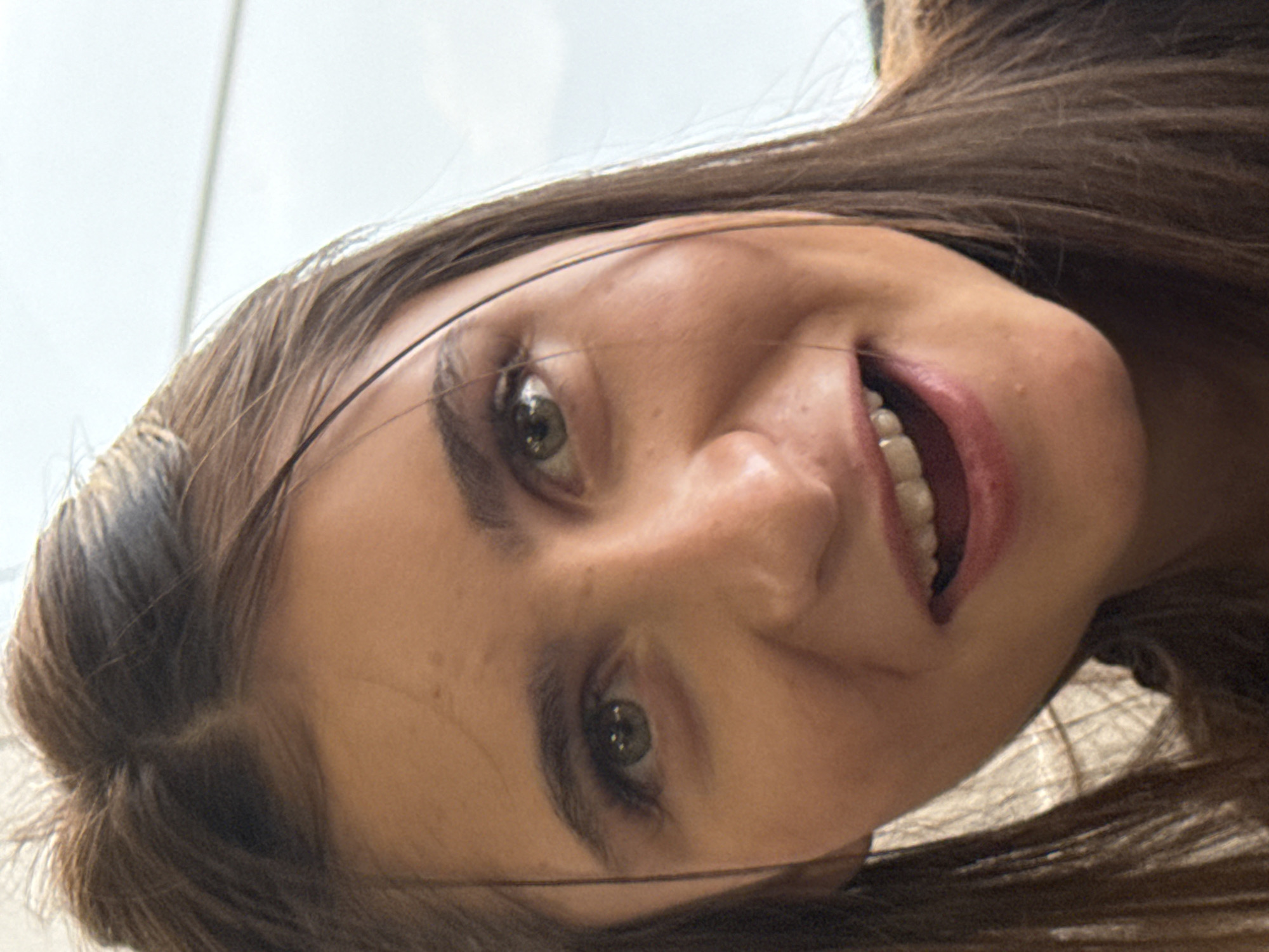 The image shows a close-up of a woman's face, angled from above and tilted. She has light skin, brown hair, and green eyes. She is smiling, revealing her teeth. The background is indistinct, suggesting a light-colored or overcast environment. No specific details indicate the location as Plovdiv, Bulgaria.