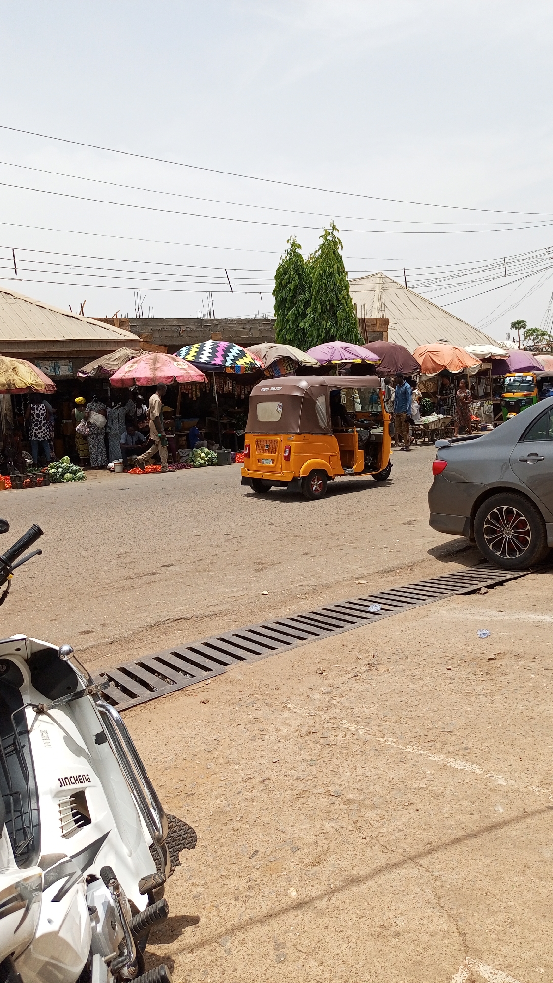 This image captures a vibrant daytime street market scene in Narayi, Nigeria. The main focus is a bustling outdoor market lining the left side of a paved road. Numerous market stalls are set up, shaded by a variety of colorful umbrellas, including solid pink, purple, orange, black, and a distinctive multi-colored zigzag pattern. Beneath these umbrellas, several people are visible, appearing to be vendors and shoppers engaging in commerce. Piles of fresh produce, notably green cabbages, are displayed on the ground in front of the stalls.
In the middle of the dusty road, an orange and brown auto-rickshaw, common in Nigeria, is positioned, facing away from the viewer. Text visible on its rear includes "RUAIY BIG STEP" and a license plate fragment "NG-480A". To the right, the rear section of a dark grey car with black and silver multi-spoke rims is partially visible. The immediate foreground on the left shows the front section of a white scooter or motorcycle, with the brand name "JINCHENG" clearly visible on its side. A long, dark metal drain grate runs across the width of the road in the foreground.
Overhead, numerous power lines crisscross against a bright, slightly overcast sky. Two tall, slender green trees stand prominently behind the market stalls on the right. The scene depicts a lively commercial area under what appears to be warm, daylight conditions, with umbrellas providing necessary shade.