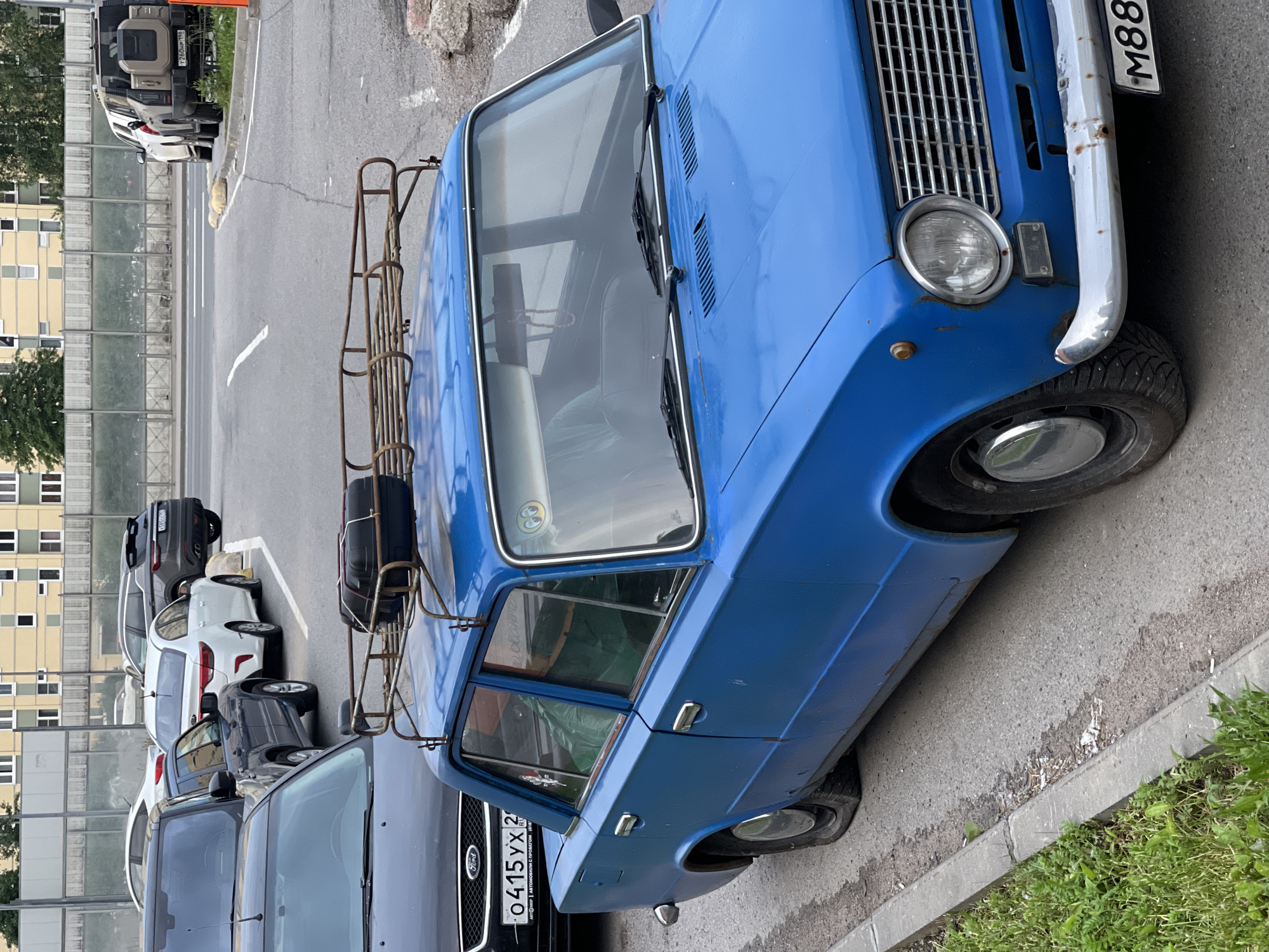A vintage blue sedan is parked on the side of a street in an urban environment. The car has a rusty roof rack with a black suitcase secured to it. The vehicle's tires appear to be flat. The car is positioned next to a curb with grass on the other side. In the background, several other vehicles are parked, including a black Ford, a white Kia, and a white SUV. Buildings and a fence are visible further in the distance. The scene suggests a parking area in a city.