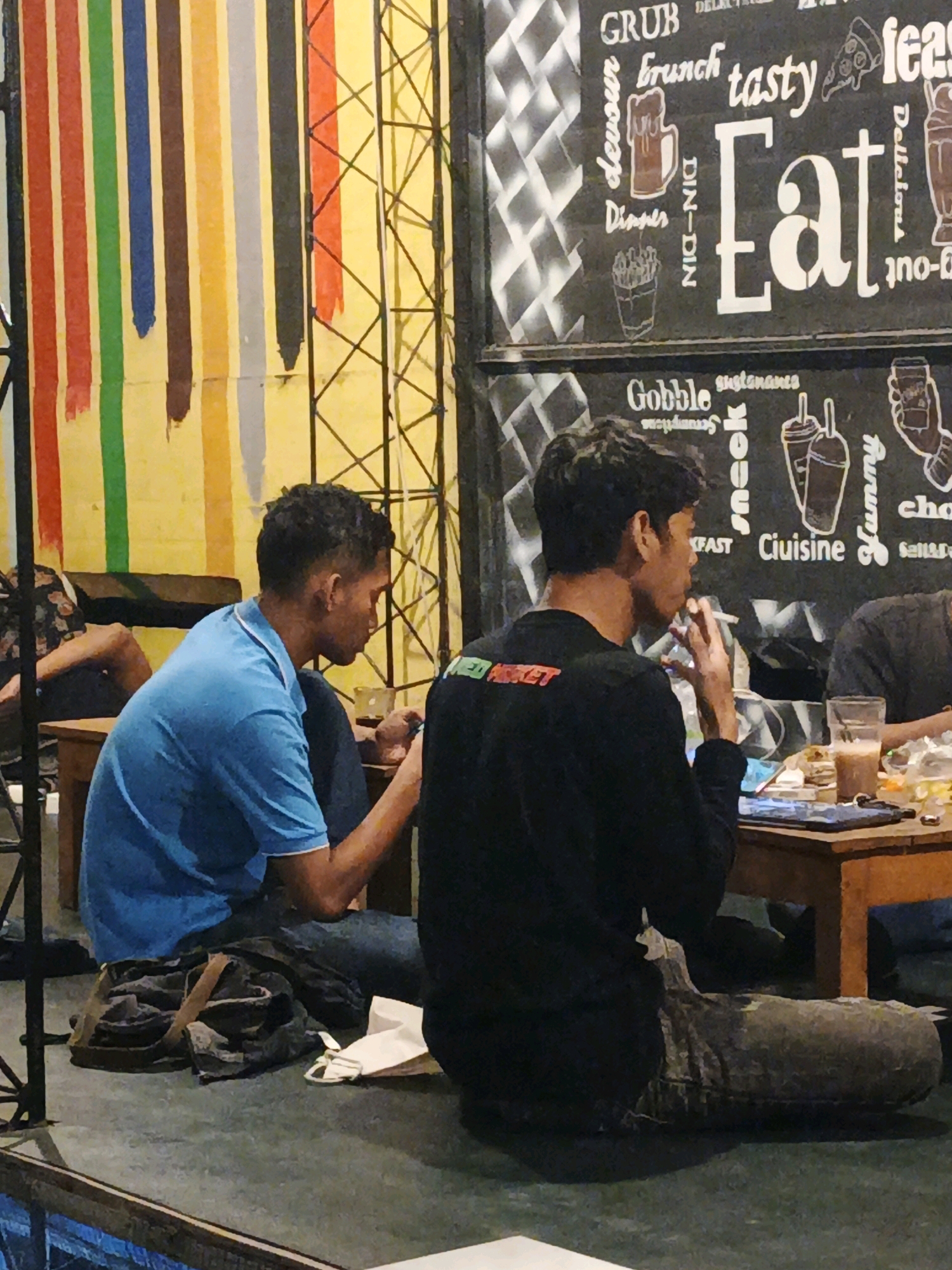 In Ketegan, Indonesia, at a cafe, two young men are seen seated at a table. One man, wearing a blue polo shirt, is looking down at his phone. The other man, in a black long-sleeved shirt with a green and red graphic on the back, is smoking a cigarette. A drink in a glass and a plate with food are on the table in front of them. The cafe's wall is decorated with colorful vertical stripes and a black chalkboard with white lettering that includes words like "Eat," "Brunch," and "Gobble." The lighting is dim, contributing to the relaxed atmosphere.