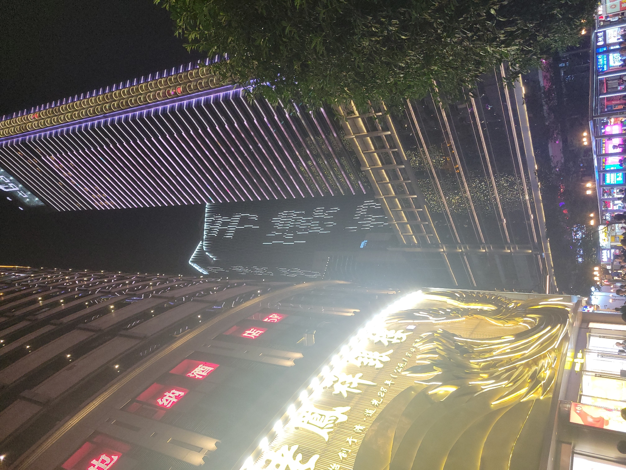 The image depicts a nighttime scene in Yuzhong District, China. There are several illuminated buildings, including one with a prominent golden dragon sculpture. Another building features rows of purple and white lights. People are present on the right side of the frame, near a brightly lit commercial area with storefronts. Tree foliage is visible in the upper portion of the image.