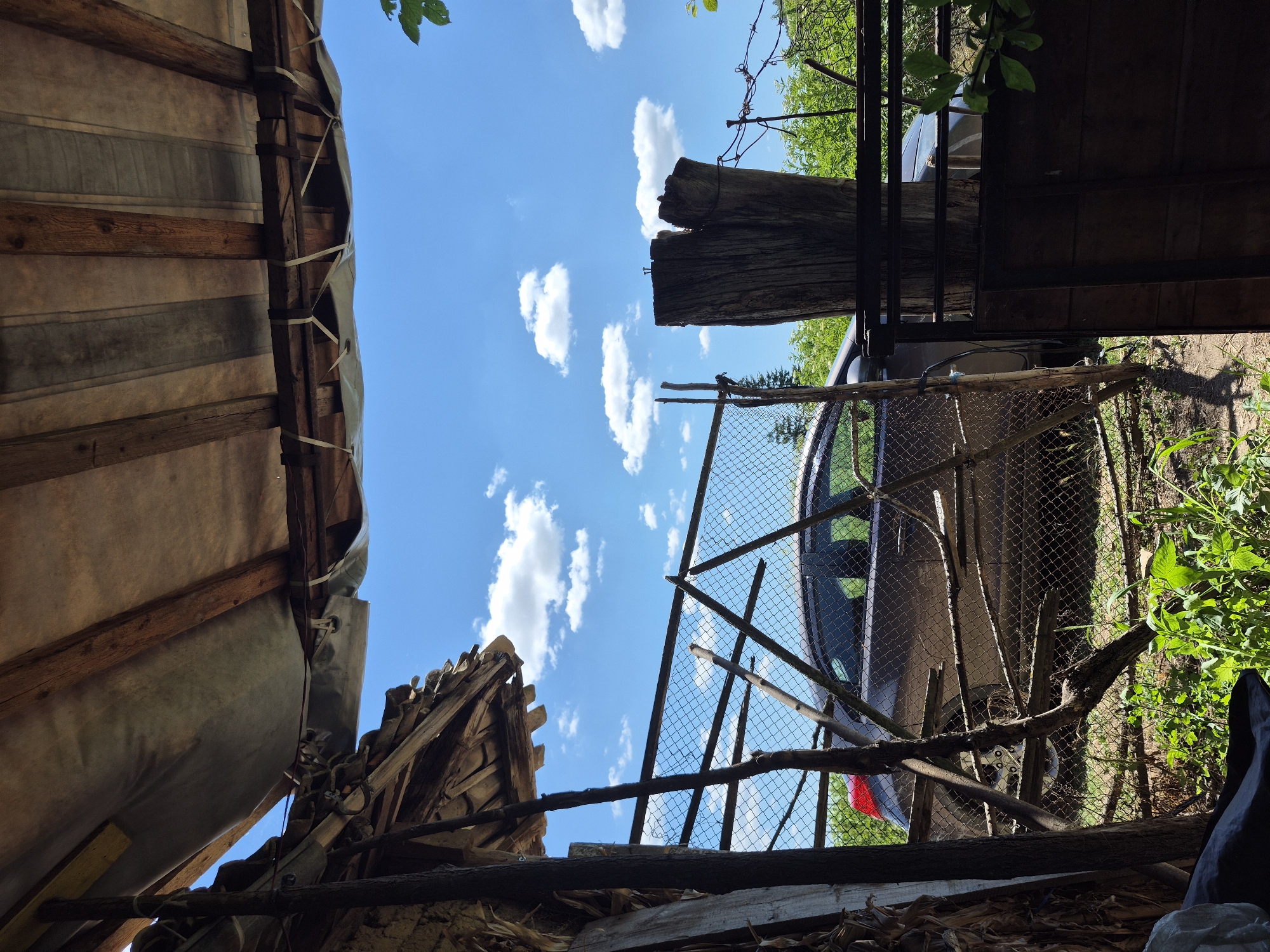 The image depicts an outdoor scene, likely a yard or rural area, under a bright blue sky with scattered white clouds. In the foreground, a makeshift shelter is visible, constructed from wooden beams and a tarp. To the left, a pile of wooden planks leans against a vertical support. A chain-link fence runs across the midground, partially obscuring a brown car. The car appears to be parked. Beyond the fence, lush green vegetation is abundant, suggesting a natural or overgrown environment. A thick, weathered wooden post stands prominently to the right of the fence, with nails visible on its surface. The overall impression is one of a rustic and somewhat unkempt setting. The specific location is identified as Sadina, Bulgaria.