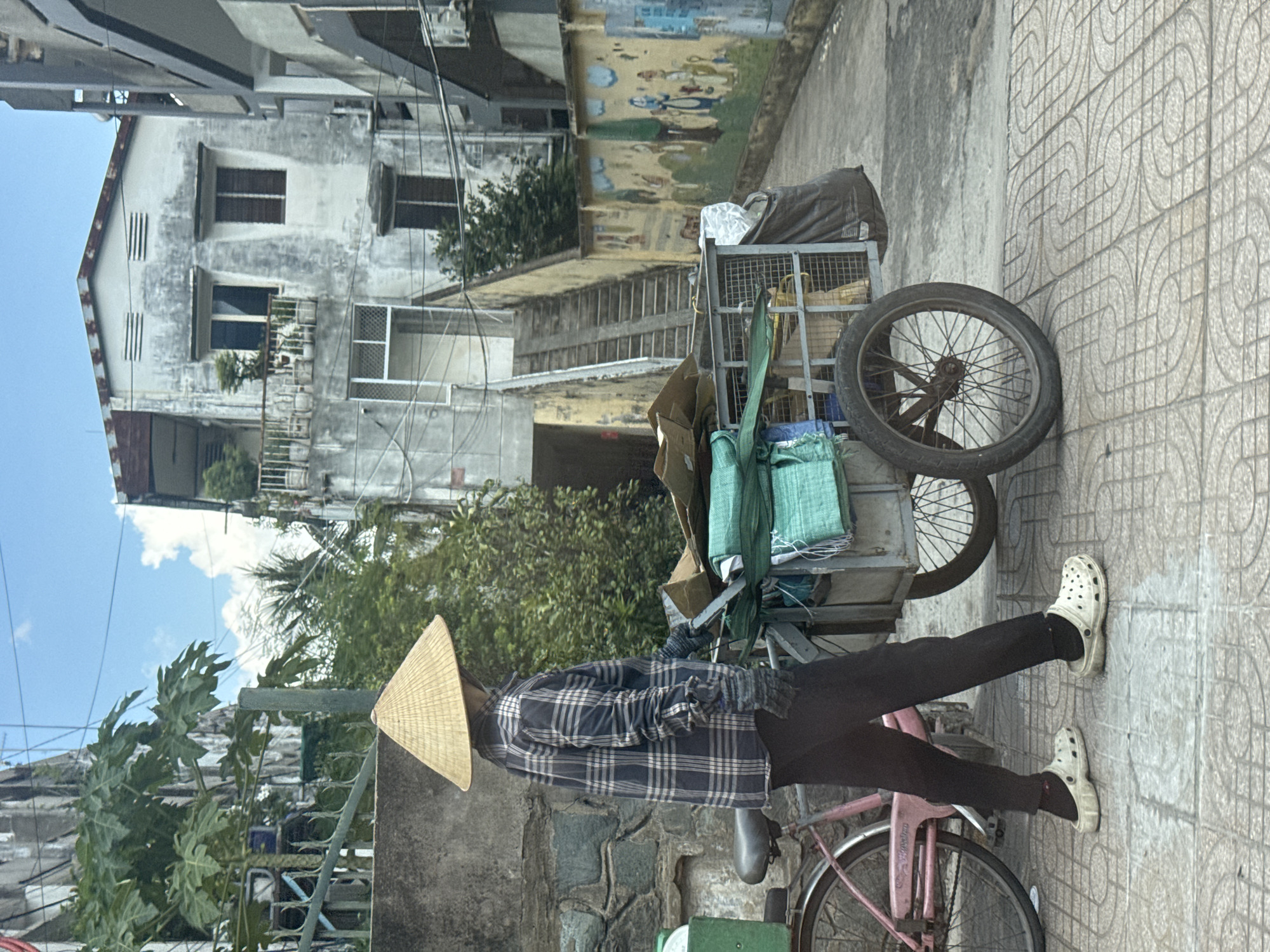 In the city of Thủ Đức, Vietnam, a person wearing a conical hat, plaid shirt, black pants, and white Crocs stands next to a pink bicycle and a metal cart. The cart, loaded with green tarps and cardboard, has two spoked wheels. The background features a weathered, multi-story building with a balcony and trees, a stone wall, and a colorful mural on a yellow wall. The ground is paved with square tiles. The scene appears to be an outdoor urban environment.