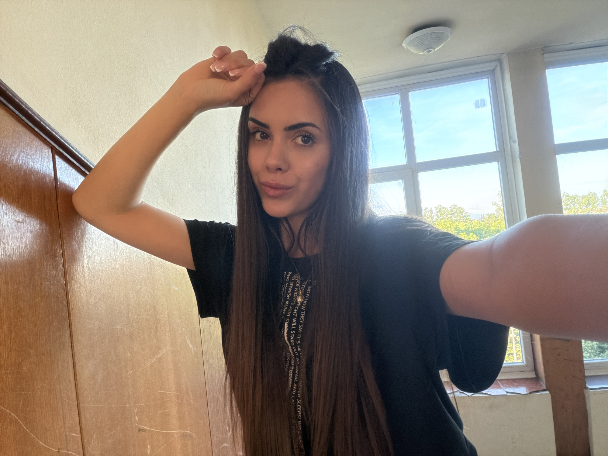 A woman is taking a selfie in what appears to be an indoor setting. She is leaning against a wooden wall with her left arm raised and hand near her head. She has long, dark hair with a small portion pulled up on top of her head. She is wearing a black t-shirt with white writing.
Behind her, there is a window divided into multiple panes, revealing a view of trees and a blue sky. The window is set within a room with light-colored walls. In the upper part of the image, there is a ceiling light fixture.