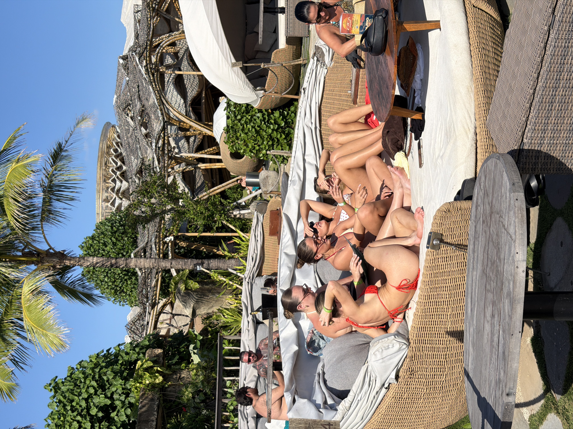 The media file depicts a group of people relaxing in an outdoor setting. Several individuals, primarily women in swimwear, are lounging on large, woven furniture. One woman, wearing sunglasses and a patterned top, is seated at a wooden table, reading a book. Others are shown in bikinis, with one in a red bikini prominently featured. Some appear to be socializing, while others are simply resting. A man with tattoos is visible in the background, also in casual attire.
The scene is set in an area with lush greenery, including palm trees and dense foliage, suggesting a tropical or subtropical environment. A distinctive architectural structure with a woven, lattice-like design is partially visible, possibly part of a resort or beach club. The bright sunlight and clear blue sky indicate it is daytime and the weather is likely warm and sunny. The overall atmosphere appears relaxed and leisurely.
No visible text is clearly discernible in the media file, although a small, dark sign with what appears to be white text is partially visible in the background foliage. The location is contextually identified as Kediri, Indonesia.