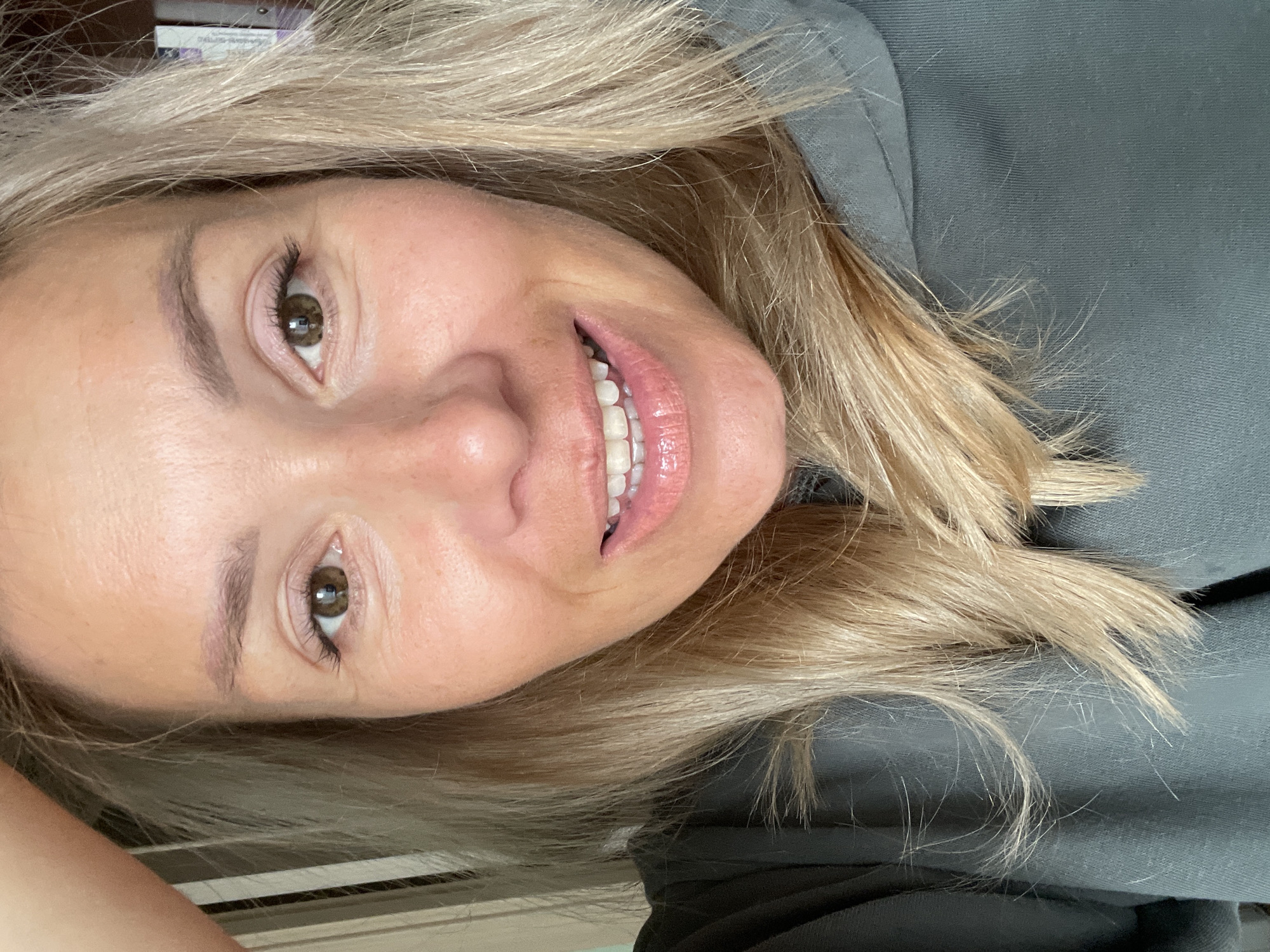 A medium shot, eye-level, selfie of a light-skinned woman with shoulder-length blonde hair and brown eyes. She is smiling and wearing a dark green hooded sweatshirt. The background is out of focus and appears to be indoors. The woman's left hand is visible in the upper left corner of the frame, resting on her head. The lighting is bright and natural.