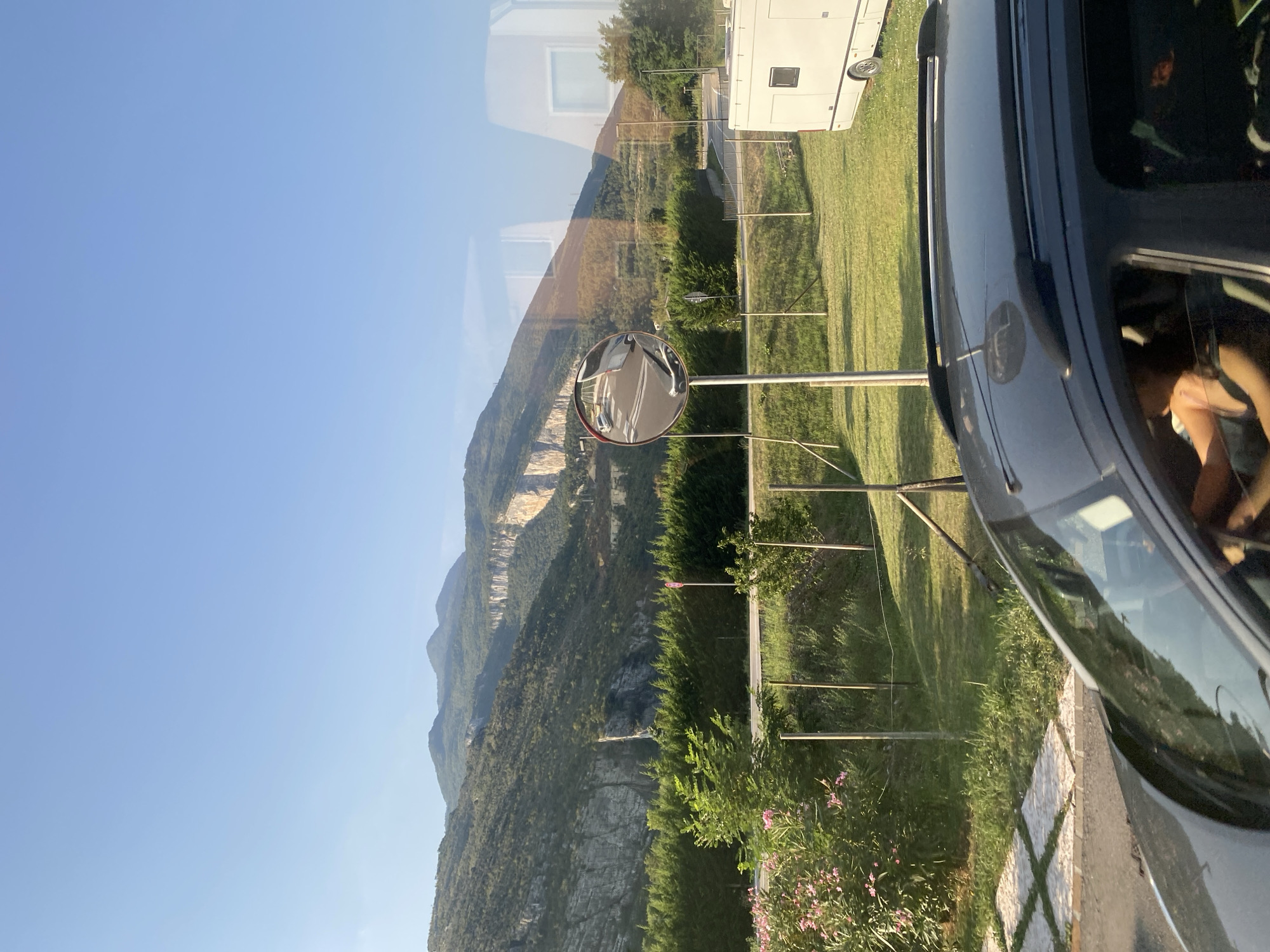 In Nago, Italy, a dark-colored SUV is parked on a street. The driver, a woman with blonde hair wearing a tank top, is visible through the driver's side window, her hands on the steering wheel. A white camper van is parked in a grassy area to the right of the SUV. In the background, a lush green hedge separates the road from the grassy area. Beyond the hedge, a winding road is visible, leading up a mountain. A convex mirror on a pole reflects cars on the road. The scene is framed by a bright blue sky and a majestic mountain range with rugged cliffs, bathed in sunlight.