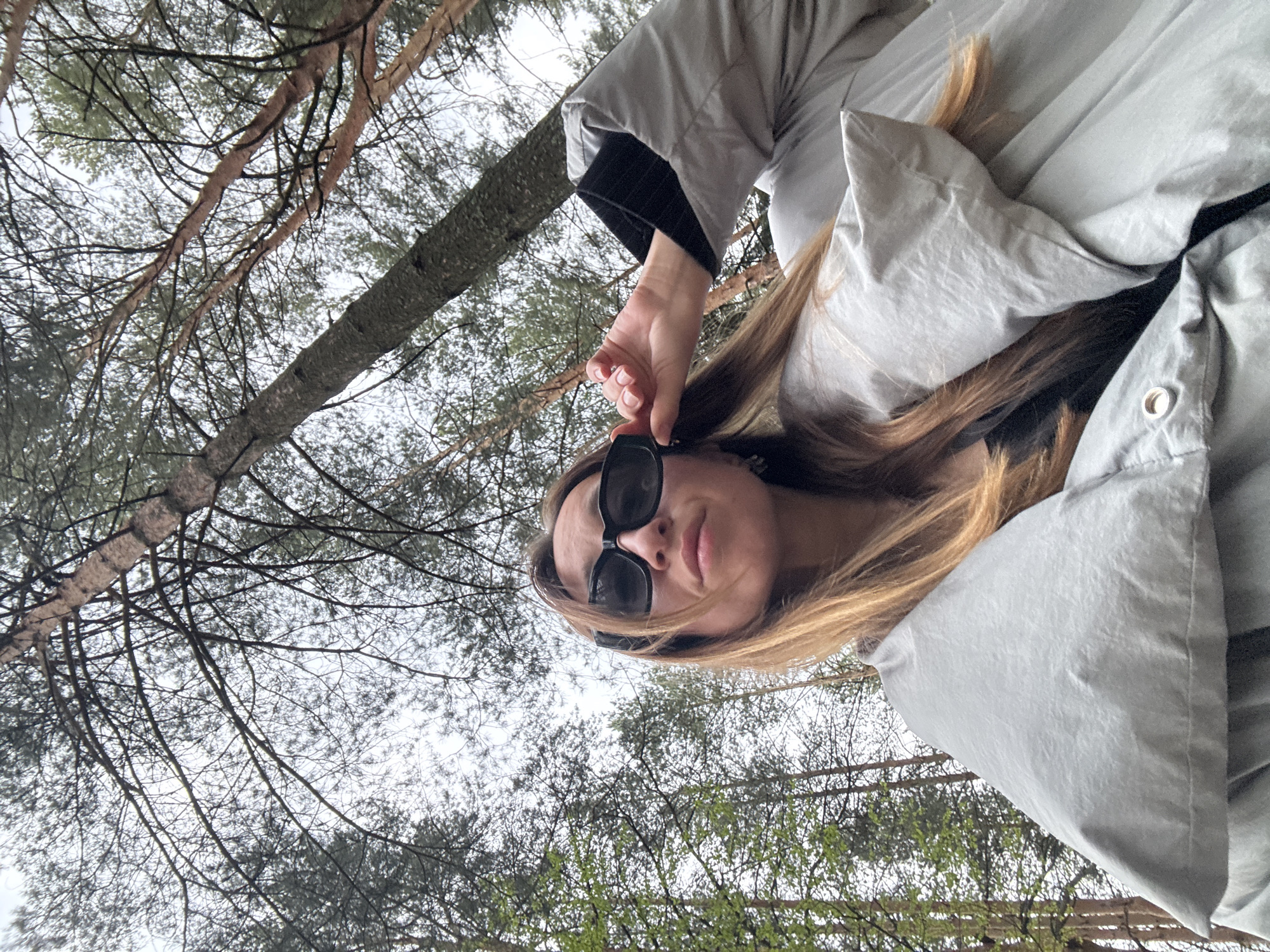 A woman wearing sunglasses and a puffy gray jacket is looking down at the camera. She is standing in a forest, as evidenced by the trees visible in the background and above her. The location is likely in Прикамский, Russia.