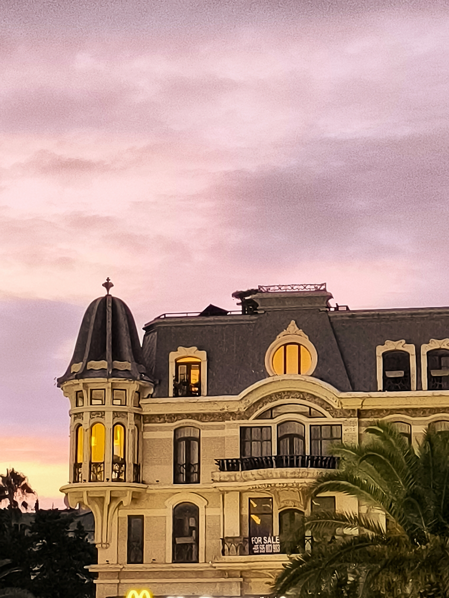 The image depicts a building with a distinctive mansard roof and a corner turret, characteristic of early 20th-century architecture, likely located in Batumi, Georgia. The sky is a soft blend of pink and purple, suggesting either sunrise or sunset. Warm light emanates from the windows of the building, indicating that the interior is illuminated. A "FOR SALE" sign is visible through a window on a lower floor. Palm fronds from a tree in the foreground partially obscure the building. The overall impression is one of serene twilight in an urban setting.