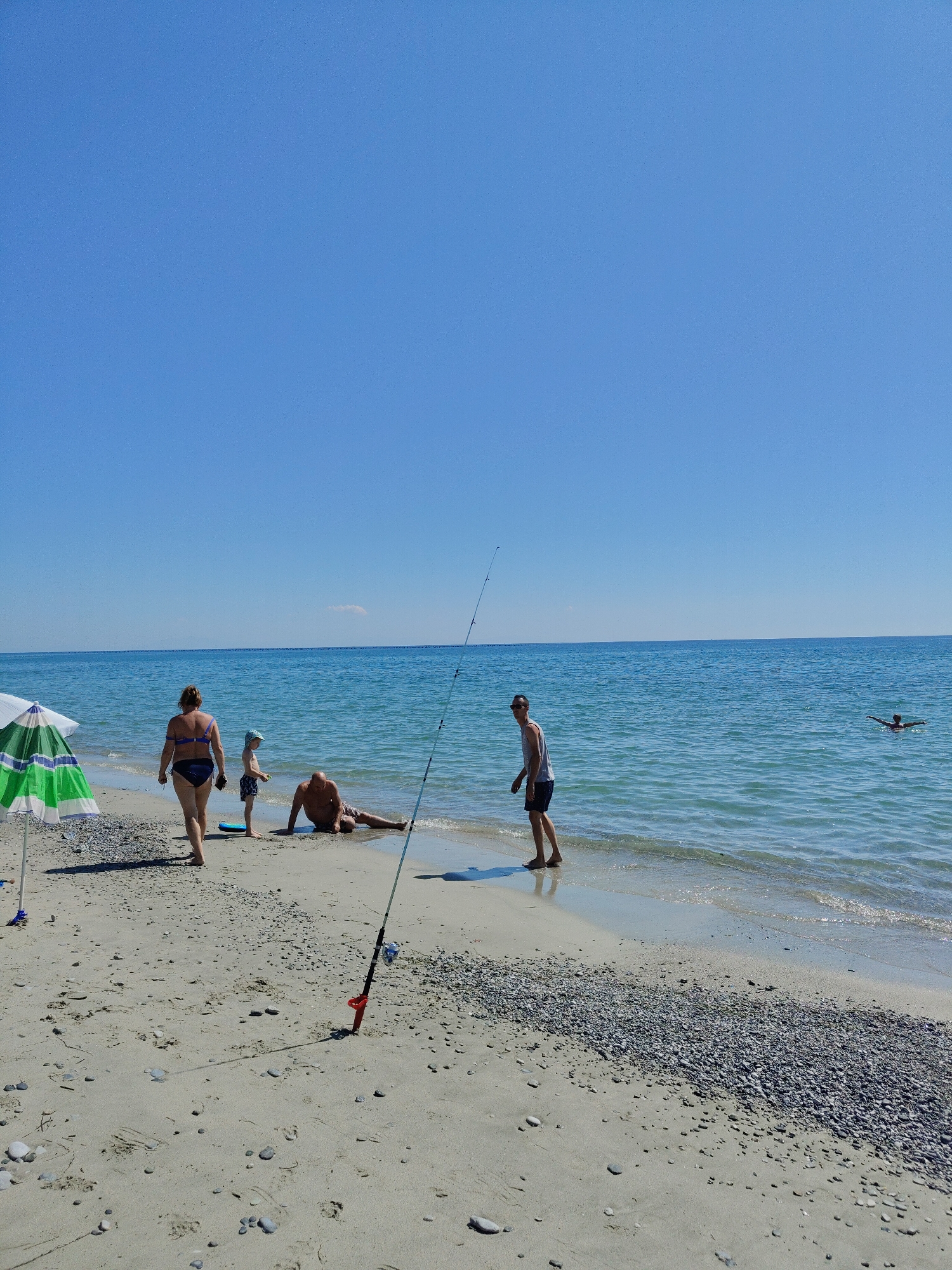 The image depicts a sunny day at a beach in Litohoro, Greece. The scene is set along the shoreline of the clear blue Aegean Sea.
In the foreground, on the sandy beach, a green and white striped beach umbrella stands to the left. Several pebbles are scattered across the sand. A fishing rod with a red handle is planted in the sand near the water's edge, with a blue and white fishing reel visible.
Several people are present. A woman in a blue bikini is walking away from the viewer, towards the water. A young child in a blue hat and patterned shorts stands near her. A man is seated on the sand, looking towards the sea. Further to the right, a man in a grey tank top and dark shorts walks along the shoreline, looking back towards the viewer. In the distant water, a person is swimming.
The sky is a clear, bright blue with no clouds visible. The overall impression is of a leisurely day at the beach.