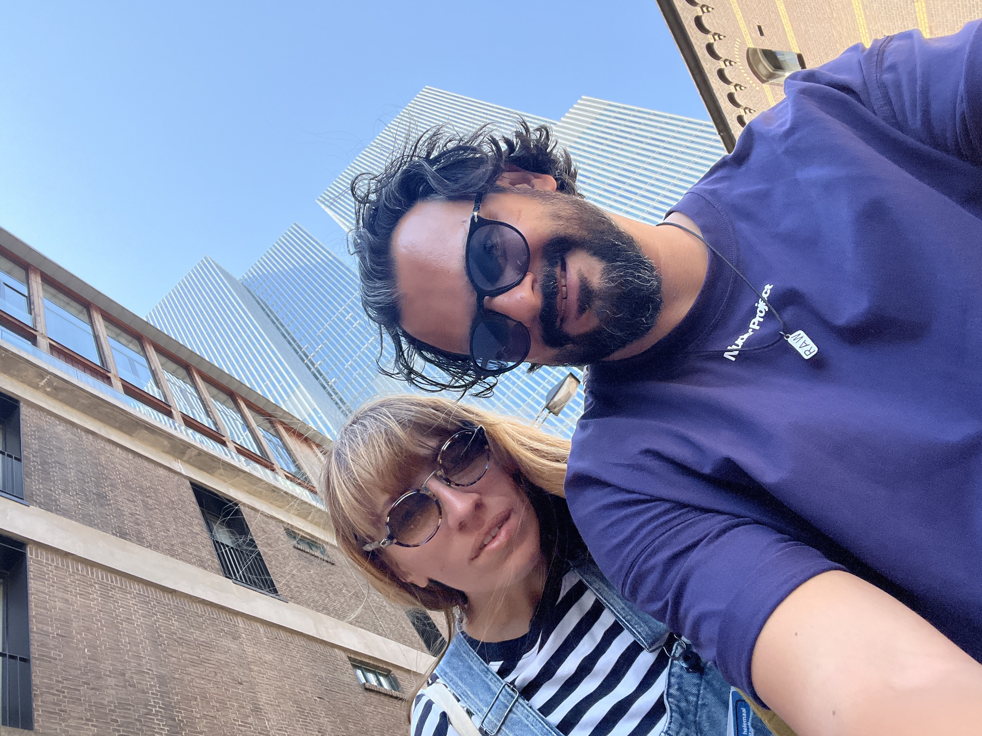 This media file is a selfie taken outdoors on a sunny day. In the foreground, a man with dark, curly hair and a beard, wearing sunglasses and a dark purple t-shirt with a small tag that reads "RAW," is looking at the camera. Next to him, a woman with blonde hair and bangs, wearing sunglasses and a striped shirt with denim overalls, is also looking at the camera.
In the background, modern, glass-paneled buildings with a distinctive striped pattern rise against a clear blue sky, suggesting an urban environment. To the left, a brick building with large windows is visible. The angle of the shot and the presence of the modern architecture strongly suggest the location is Rotterdam, Netherlands. The overall mood appears casual and happy, as if the subjects are enjoying a day out. The lighting indicates it's daytime.