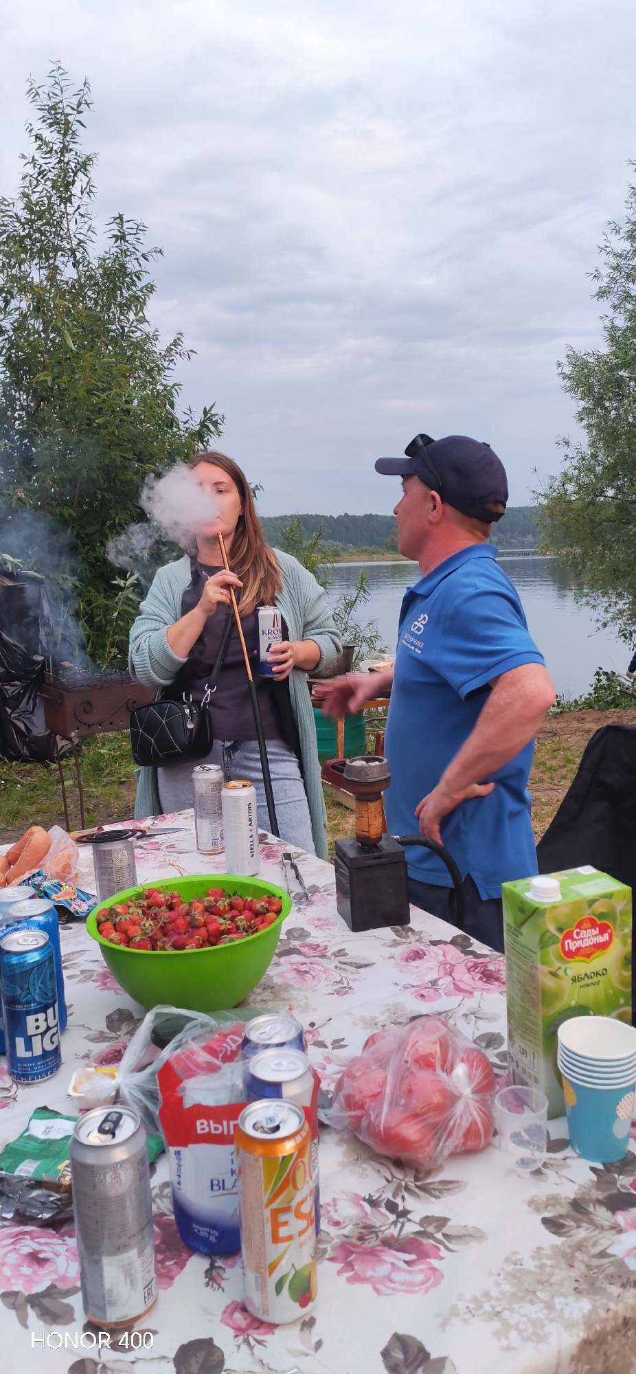 The image depicts a casual outdoor gathering near a body of water, likely a river or lake, in Noviy, Russia. A woman is smoking a hookah, exhaling a cloud of smoke. She is wearing a light blue cardigan over a dark top and blue jeans, with a black crossbody bag. She holds a can of "Kronenbourg 1664 Blanc" in her right hand. Beside her, a man in a blue polo shirt and a dark baseball cap is facing her, gesturing with his right hand, and appears to be speaking. On the table in front of them, a green bowl is filled with strawberries. There are several open cans of beer, including "Bud Light" and "Stella Artois," and a carton of apple juice labeled "Сады Придонья" (Sady Pridonya). A plastic bag filled with tomatoes is also visible on the table, along with a stack of disposable cups. A charcoal grill is present to the left of the woman, with smoke emanating from it. The background shows lush green trees and a hazy sky, characteristic of a temperate climate. The overall scene suggests a relaxed social event, possibly a picnic or barbecue, by the water.