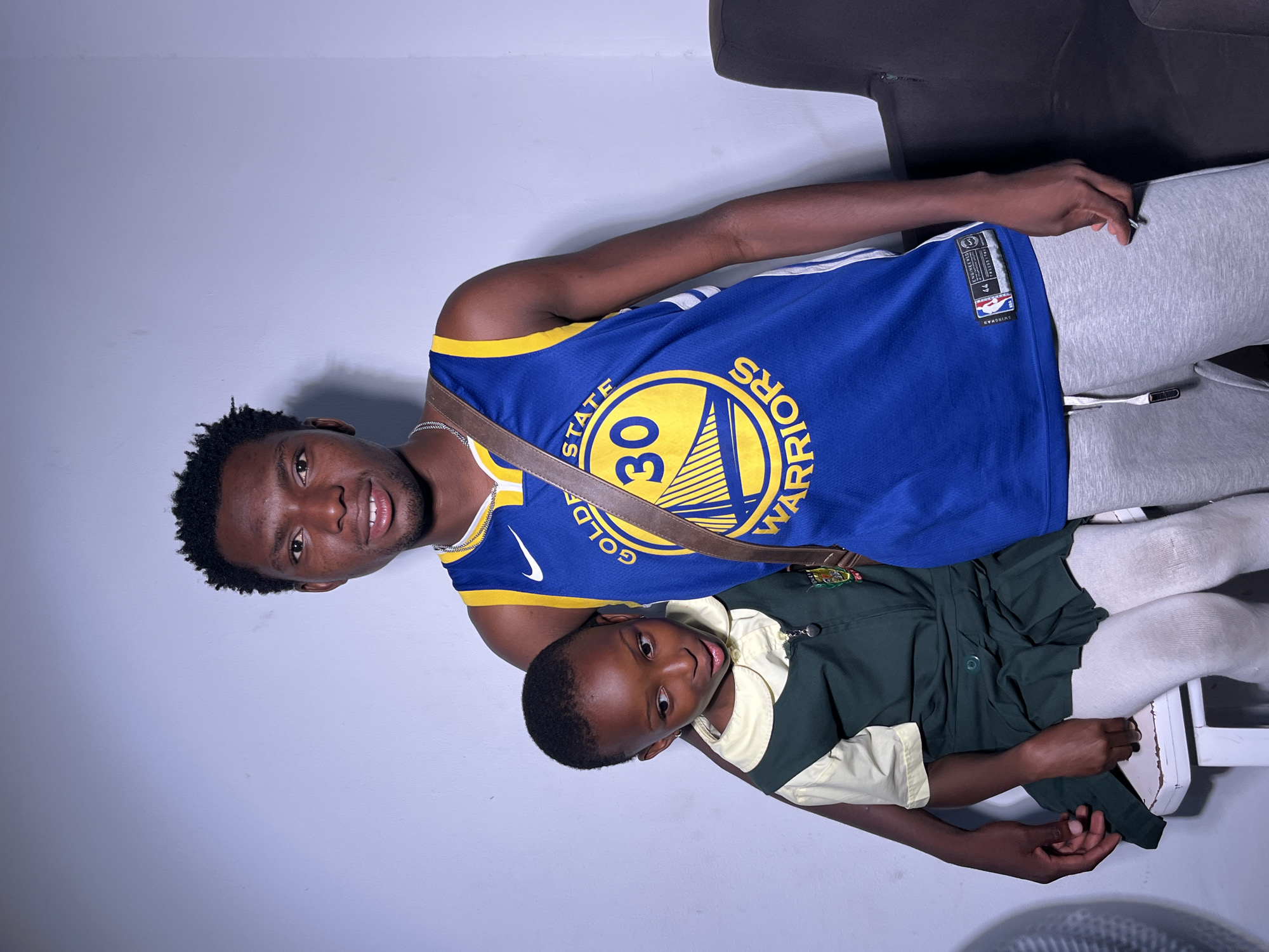 A young Black male, wearing a blue and yellow Golden State Warriors basketball jersey with the number 30 and a tan crossbody bag, stands and wraps his arm around a young Black female. The female, wearing a dark green and light yellow school uniform with white tights, sits beside him. They are positioned in front of a plain, light-colored wall. The environment appears to be indoors with a darker area to the right of the frame.