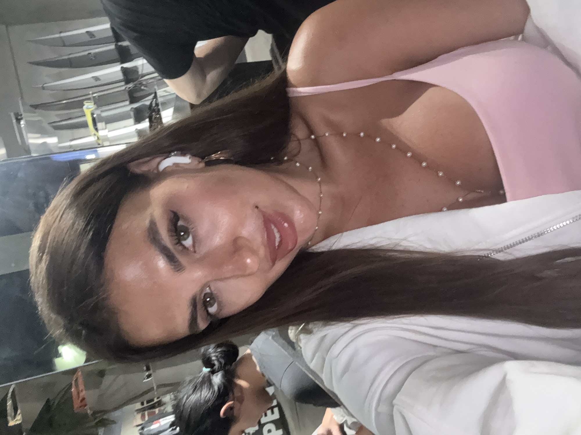 A woman with long brown hair and an earbud in her ear is the main subject of the image. She is wearing a pink top and a white jacket. A pearl necklace is visible around her neck. Behind her is a blurred view of what appears to be surfboards displayed on a rack, suggesting a surf shop or rental location. Another person with their hair in a bun is partially visible in the foreground. The background indicates the location may be North Kuta, Indonesia, given the presence of surfboards, which are common in that area.