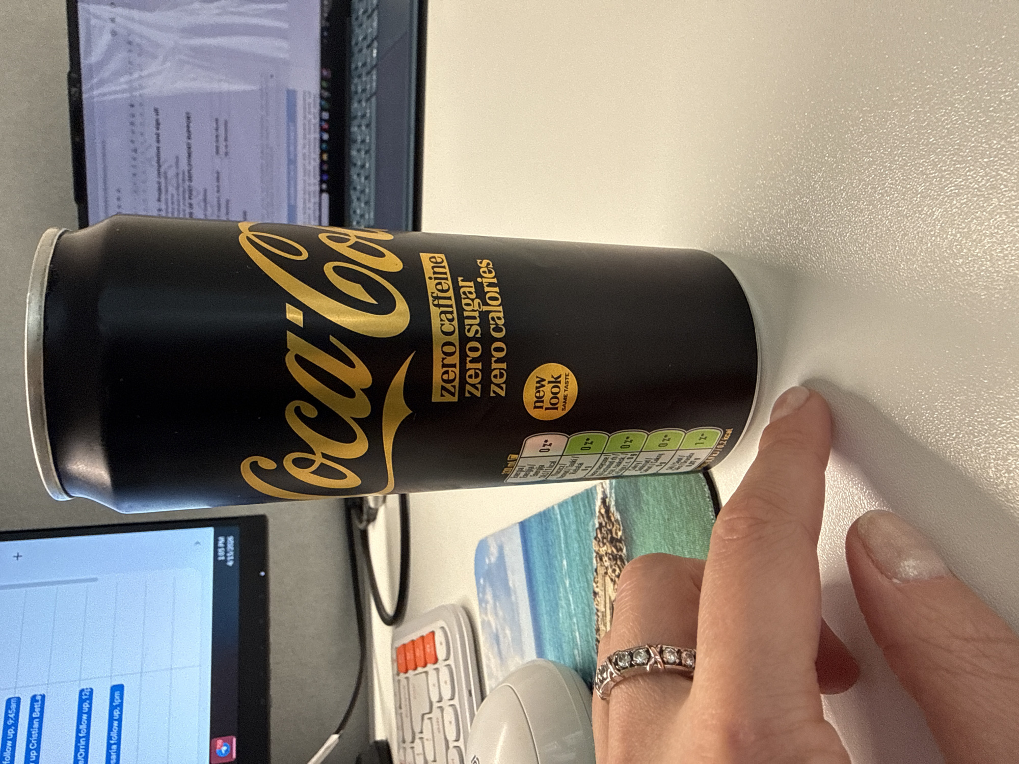 The image shows a can of Coca-Cola Zero Sugar Zero Caffeine in black and gold packaging lying on a white surface. To the left are portions of two computer monitors, a keyboard, and a mouse, suggesting an office environment. A hand with a ring on the finger is visible near the can. The OCR indicates text related to appointments or reminders and nutritional information from the can. The image does not contain enough information to determine that the image was taken in Riga, Latvia.