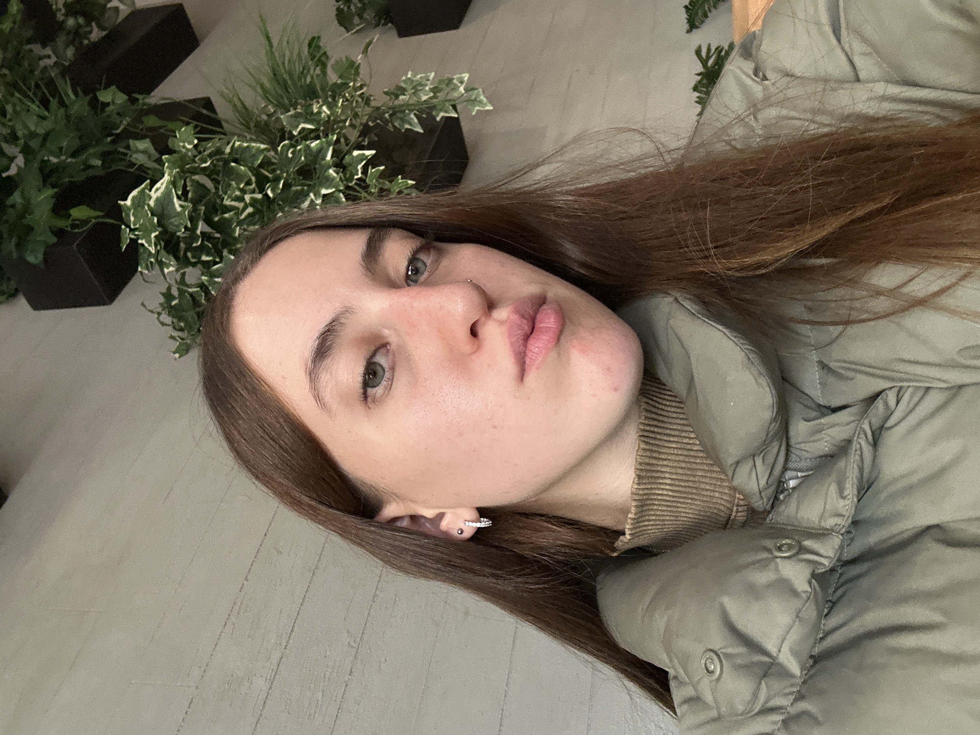 A woman with long brown hair and light skin is taking a selfie. She is wearing a green puffer jacket and a brown ribbed sweater. She has green eyes and is making a slight pout. The background includes a grey wall and several potted plants with green and white leaves. The location cannot be determined to be Huixquilucan de Degollado, Mexico.
