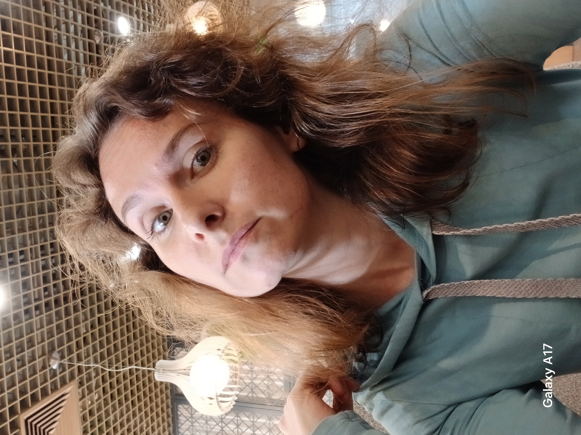 The image presents a woman, likely in her mid-to-late 30s or early 40s, captured indoors in Yekaterinburg, Russia. Her head is tilted to her right (viewer's left), and her light-colored eyes, appearing green or hazel, gaze slightly upward towards the camera with a neutral expression. Her hair is light brown to auburn, voluminous, and appears somewhat disheveled, extending past her shoulders. She is dressed in a teal or seafoam green hooded sweatshirt featuring visible grey, textured drawstrings at the neck. Her left hand (viewer's right) is partially visible, with fingers gently positioned within her hair on the left side of her head. The background consists of a light-colored, square-grid ceiling structure. A distinct white, decorative pendant light fixture, housing an illuminated bulb, hangs prominently to the left of the woman's head. Additional blurred light sources are visible in the background. The text "Galaxy A17" is imprinted in the lower-left corner of the image.