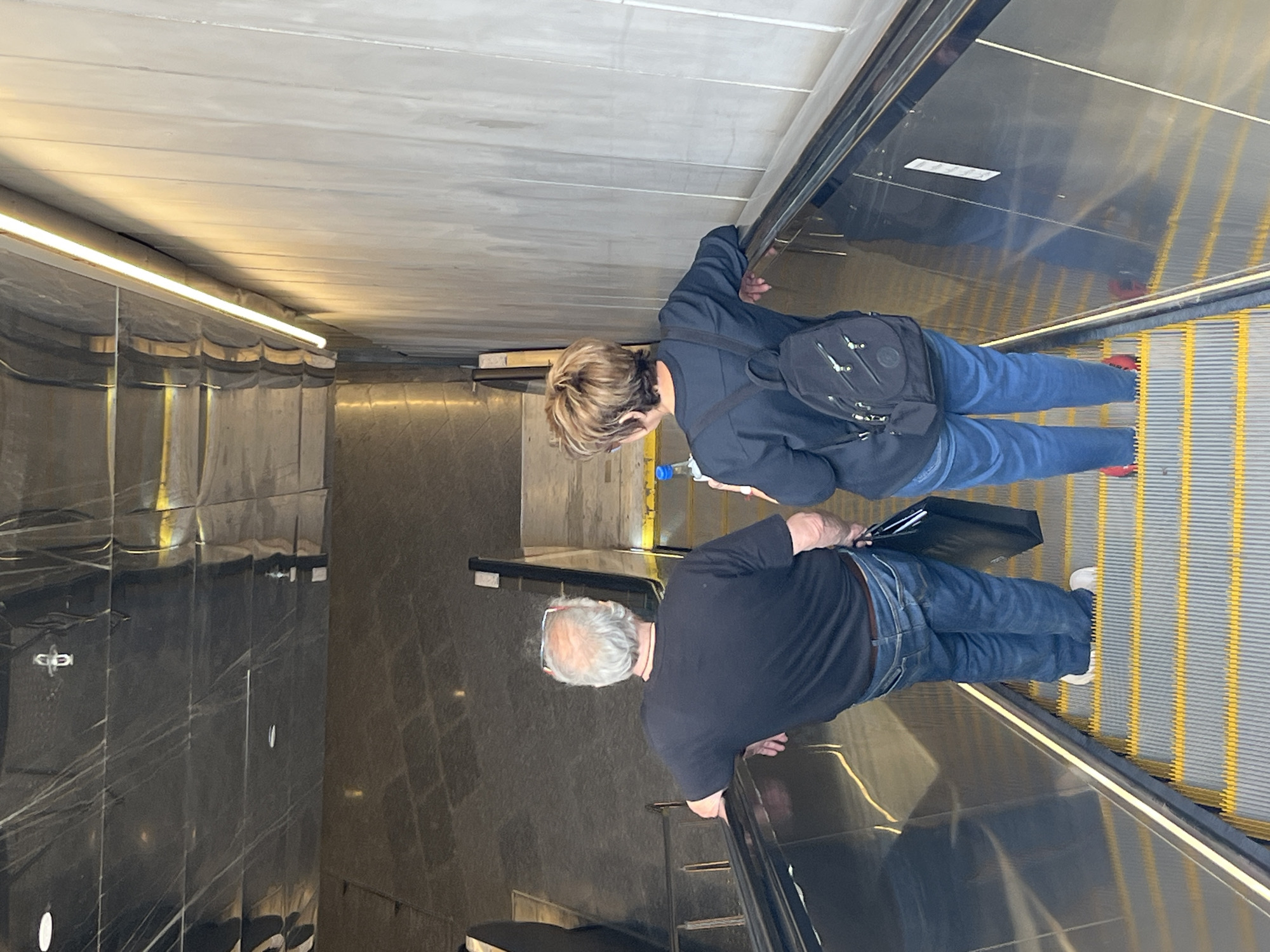 The image shows two people on an escalator. A woman with short blonde hair, wearing a dark blue jacket, jeans, and red sneakers, is carrying a black backpack. A man with grey hair, wearing a black shirt, jeans, and white sneakers, is holding onto a black briefcase. They are riding the escalator down. The environment features reflective walls and a fluorescent light overhead. The image appears to be taken in a modern, possibly underground or transportation-related setting. No specific information is available in the image to identify the location as Tel-Aviv, Israel.