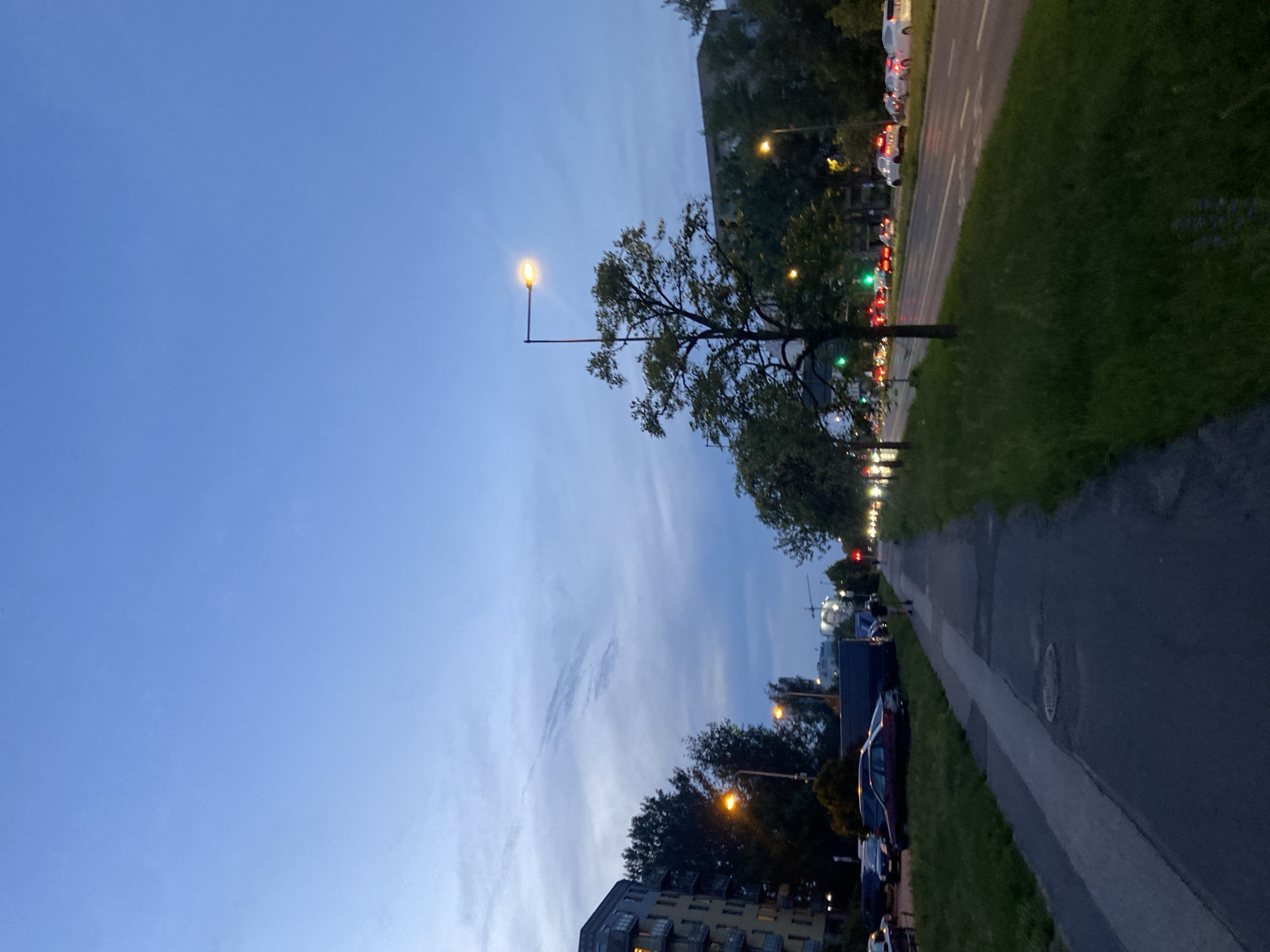 The image captures a scene on a street in Warsaw, Poland, likely during dusk or early evening, as evidenced by the clear blue sky transitioning into twilight and the illuminated streetlights and vehicle headlights. A paved sidewalk runs along the left side of the frame, bordered by a strip of green grass. Parked cars are visible along the sidewalk, adjacent to a multi-story building.
Across the street, a roadway with multiple lanes is visible, with traffic flow indicated by the headlights and taillights of moving vehicles. Streetlights illuminate the road, and trees line the right side of the roadway, their leaves appearing dark against the twilight sky. In the distance, buildings and structures can be seen, including what appears to be a construction crane. A single person is walking on the sidewalk in the mid-ground, heading away from the camera. The overall environment suggests an urban setting within Warsaw, characterized by residential buildings, roadways, and pedestrian infrastructure.